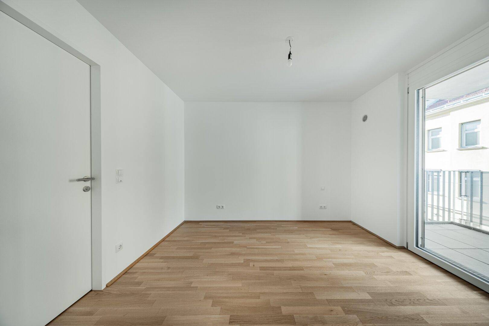 Heller, leerer Raum mit Parkettboden und großem Fenster, das viel Licht spendet.