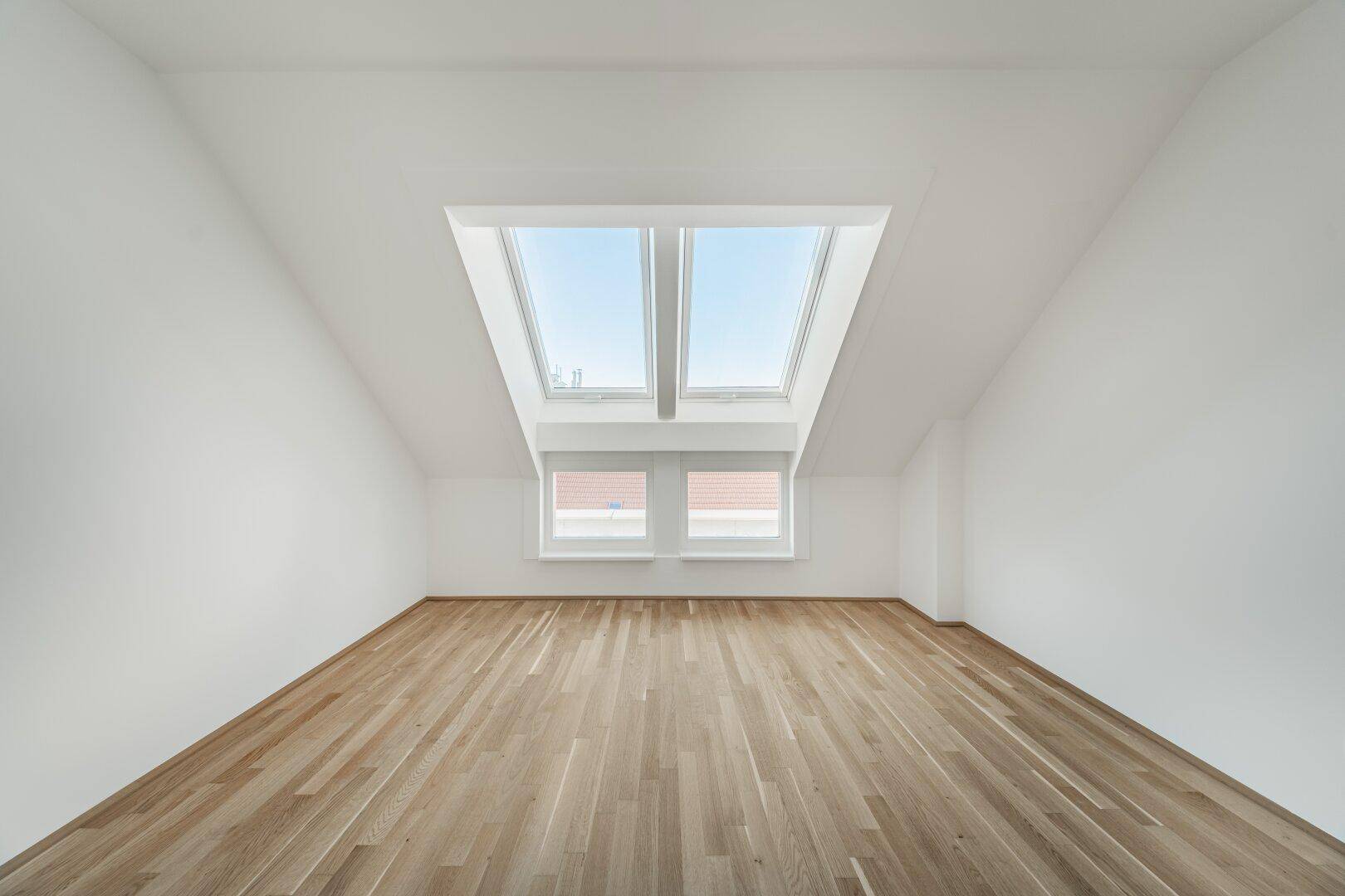 Geräumiges Zimmer im Dachgeschoss mit Holzboden, schrägen Wänden und zwei großen Dachfenstern.