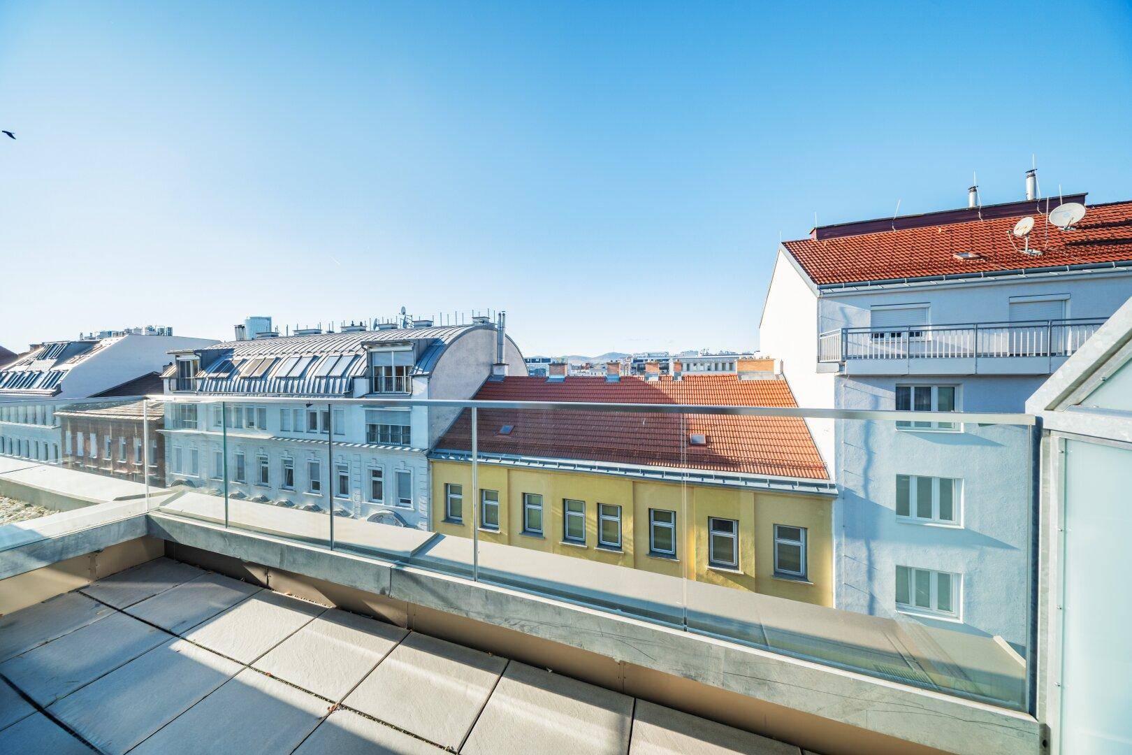 Sonnige Dachterrasse mit Glasgeländer und Blick über die Dächer der Stadt.