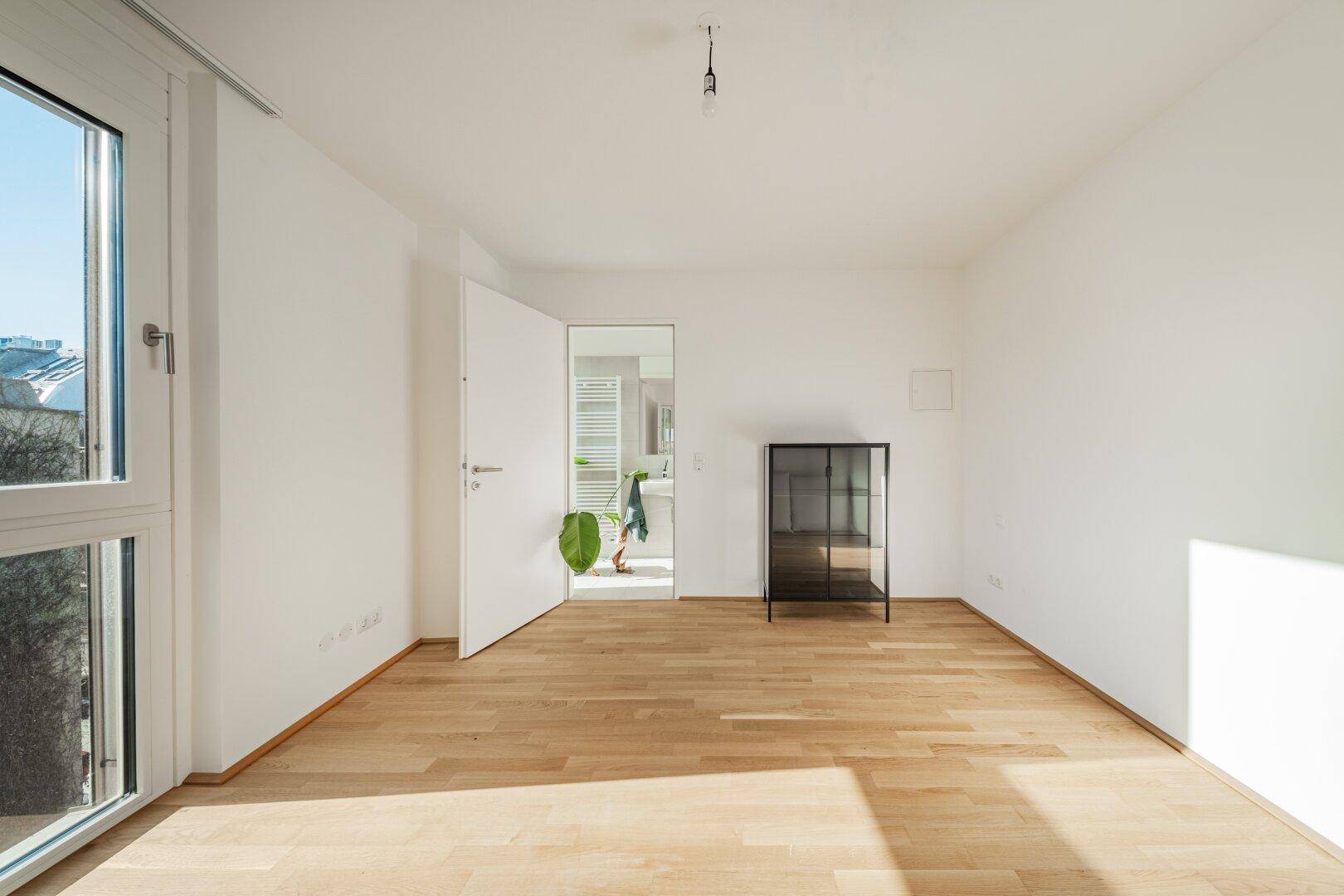Leeres Schlafzimmer mit Holzboden und großen Fenstern, die viel Licht hereinlassen.