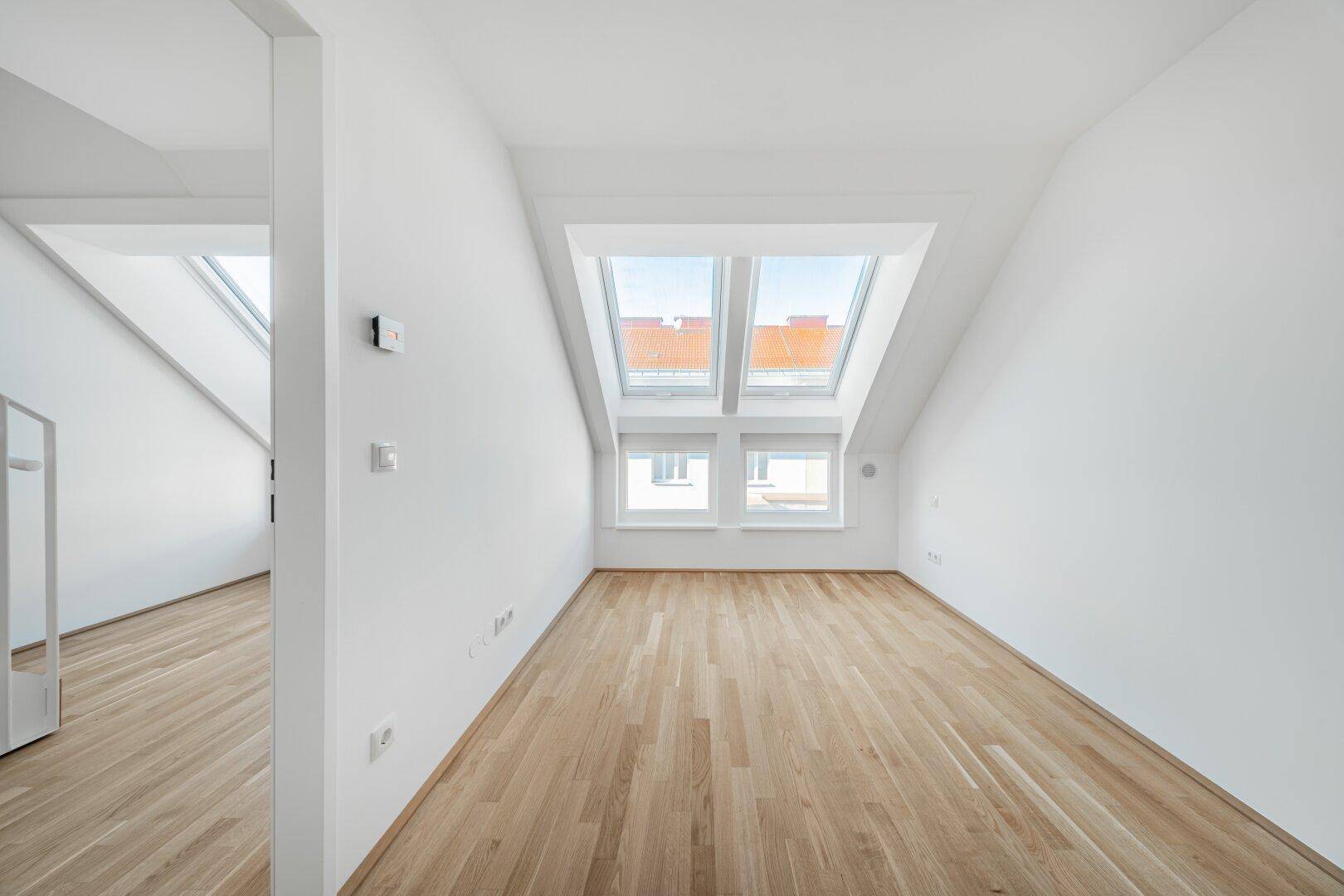 Lichtdurchfluteter Raum im Dachgeschoss mit Parkettboden, schrägen Wänden und großen Fenstern.