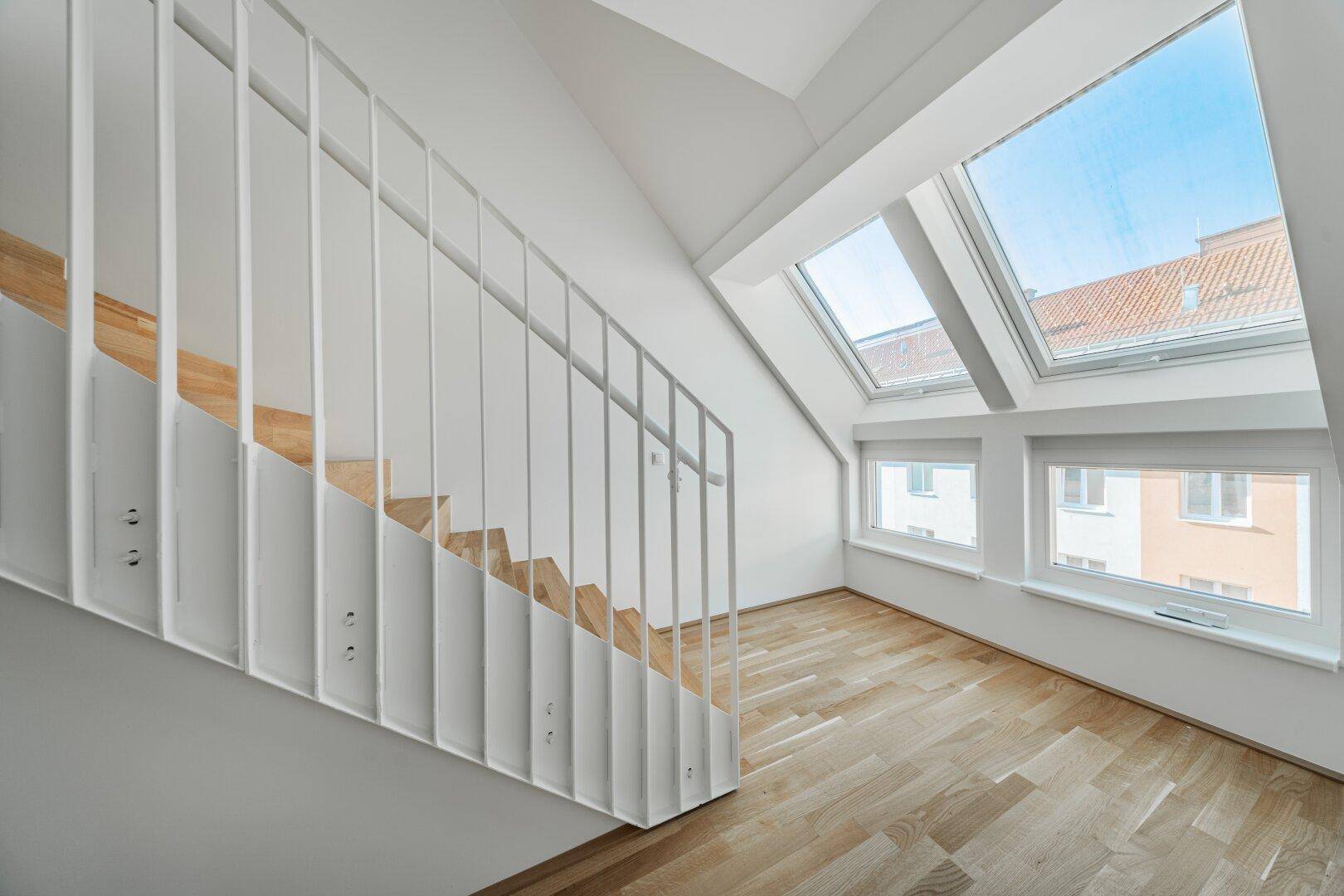 Helle Holztreppe mit modernem Geländer führt zu einem lichtdurchfluteten Dachgeschoss mit Parkettboden.