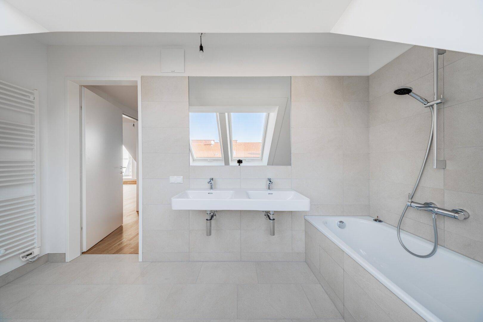 Helles Badezimmer mit Badewanne, Doppelwaschbecken und beheiztem Handtuchhalter, angrenzend an einen Wohnraum.