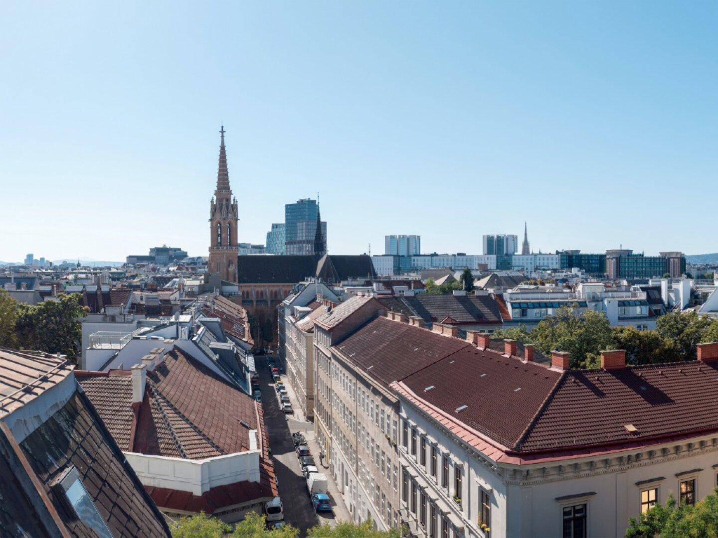 Weitläufiger Stadtblick mit einer Kirche, historischen Gebäuden und modernen Hochhäusern am Horizont.