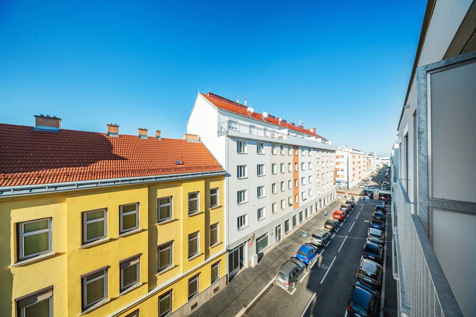 Sonniger Blick von einem Balkon auf eine belebte Straße mit parkenden Autos und Gebäuden.
