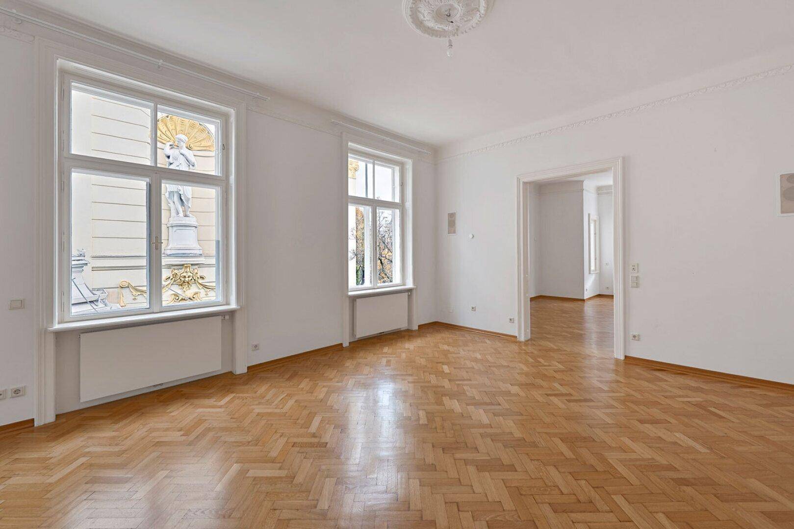 Helles Zimmer mit Fischgrätparkett, hohen Decken und Blick auf die reich verzierte historische Fassade.