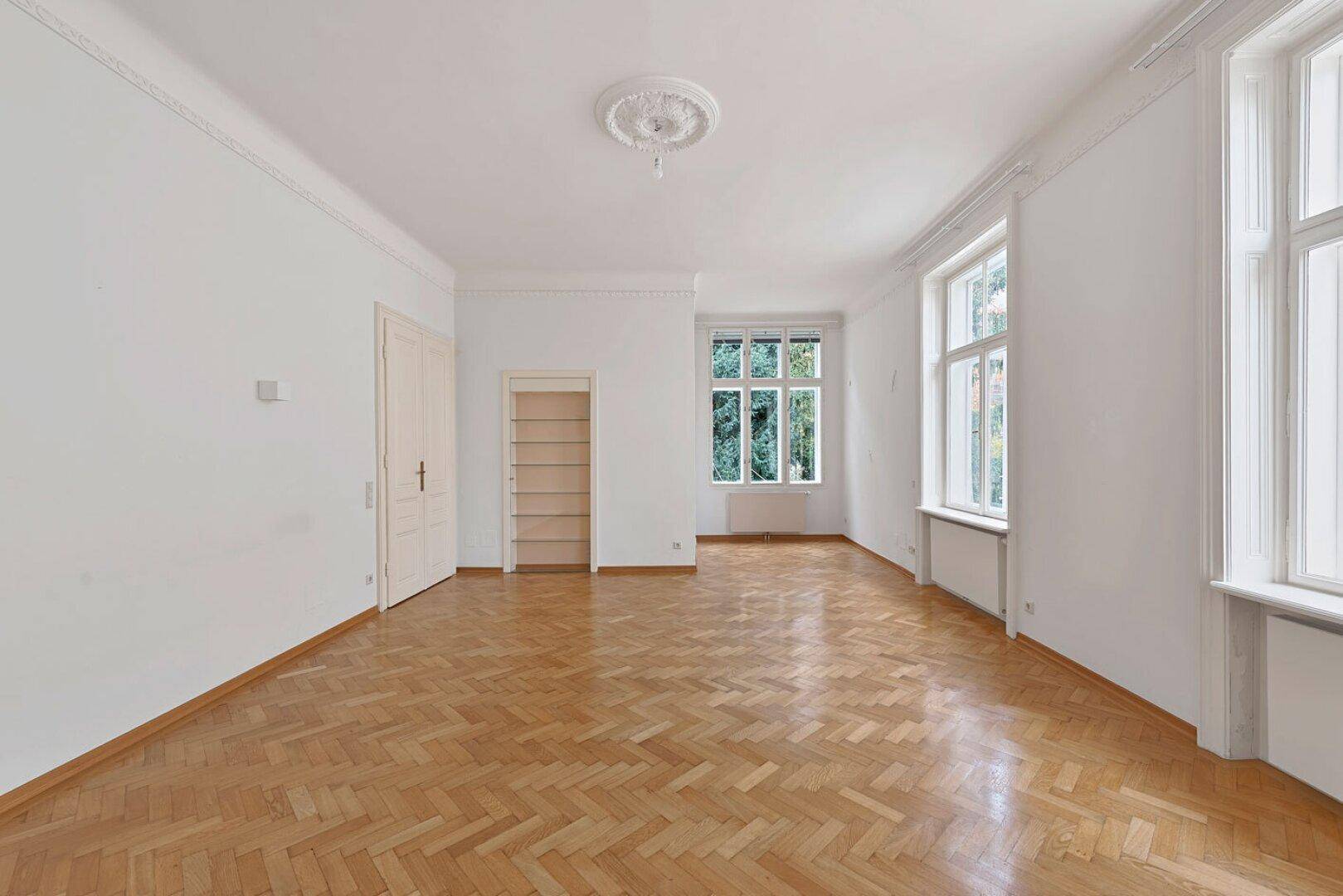 Großes, helles Zimmer mit Fischgrätparkett, hohen Decken und Blick ins Grüne durch hohe Fenster.