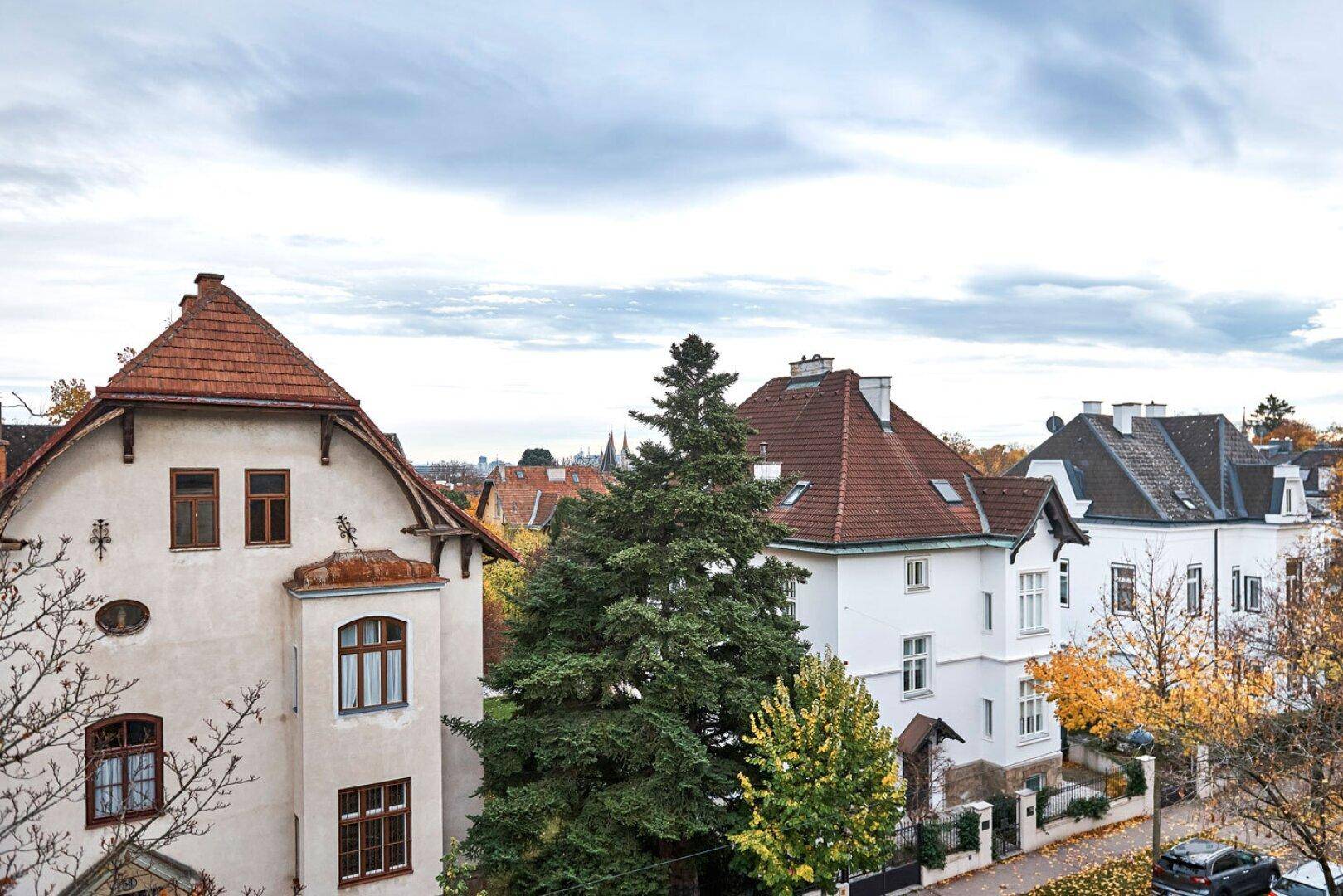 Panoramablick über eine Wohngegend mit verschiedenen Dächern und herbstlicher Baumlandschaft unter bewölktem Himmel.