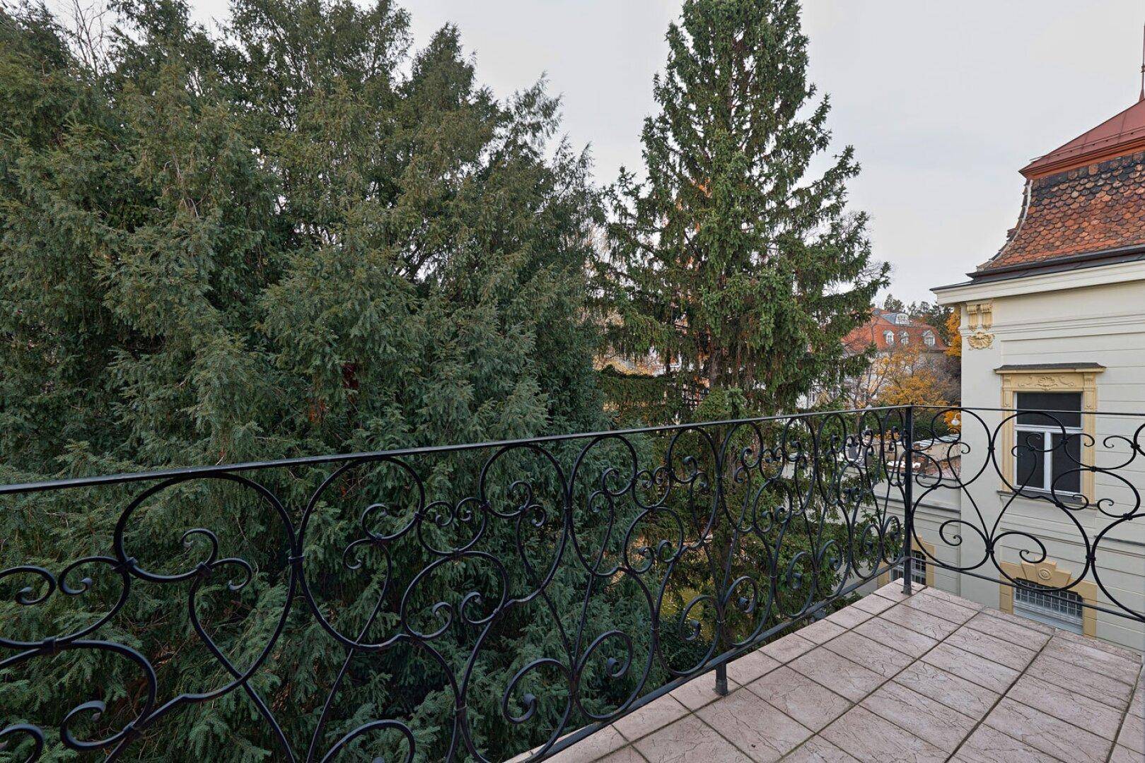 Balkon mit schmiedeeisernem Geländer und Blick auf üppiges Grün und hohe Bäume, ideal zum Entspannen.