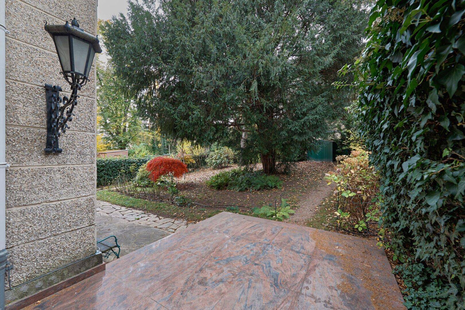 Kleine Terrasse mit Steinboden und Blick auf den gepflegten Garten mit altem Baumbestand und viel Grün.