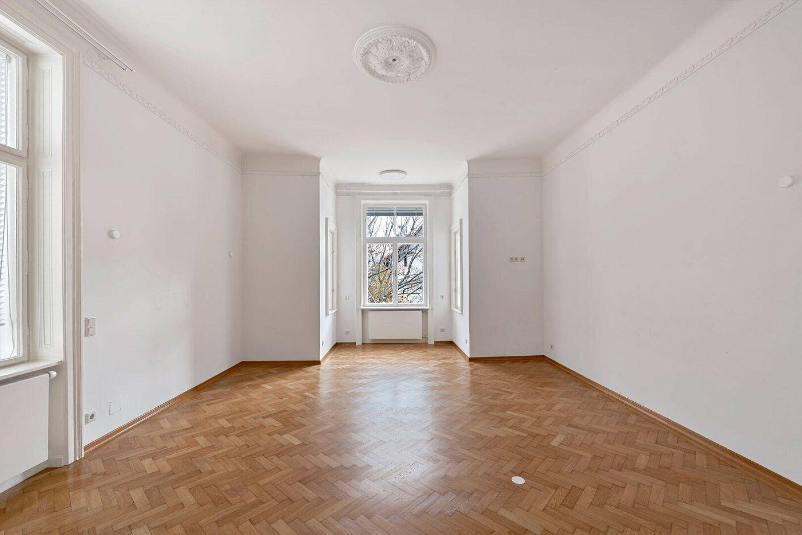 Geräumiges, helles Zimmer mit Fischgrätparkett, hohen Decken und kunstvollen Stuckelementen an der Decke.