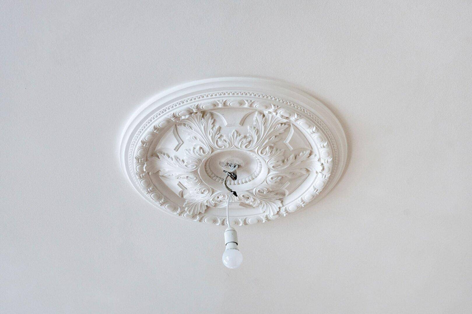 Detailaufnahme einer kunstvollen Stuckrosette an der Decke mit freiliegender Glühbirne, typisch für Altbauten.