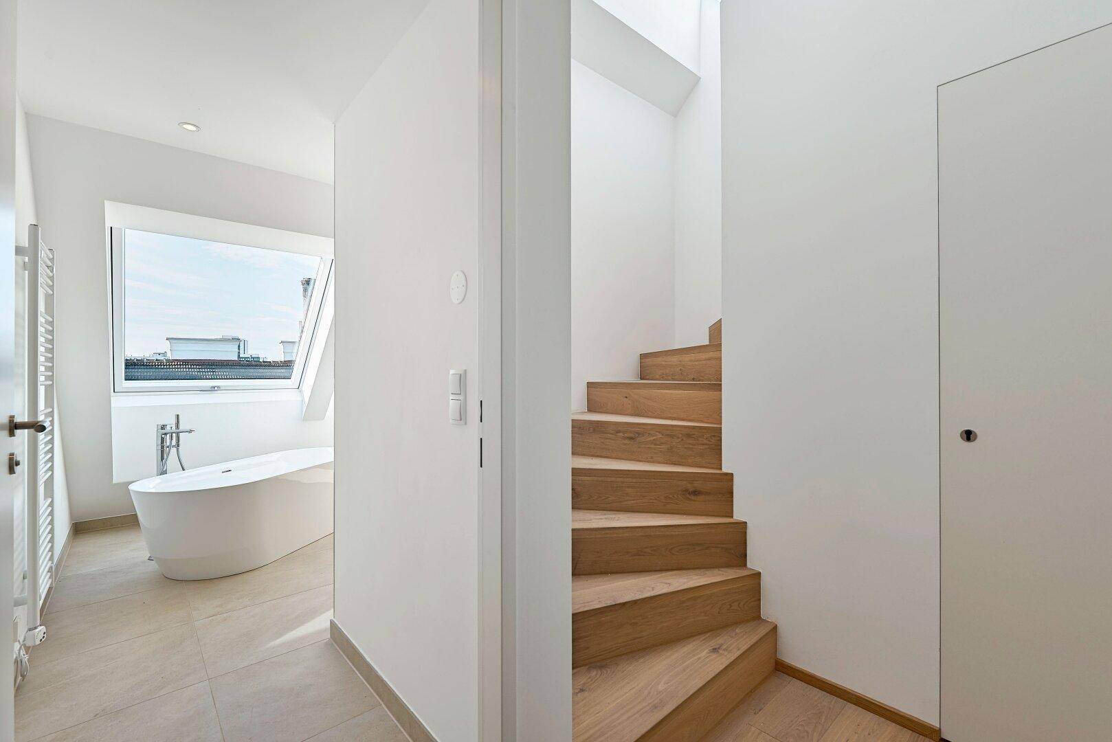 Heller Flur mit Holztreppe, die in die obere Etage führt, und Blick in ein Badezimmer mit Badewanne.