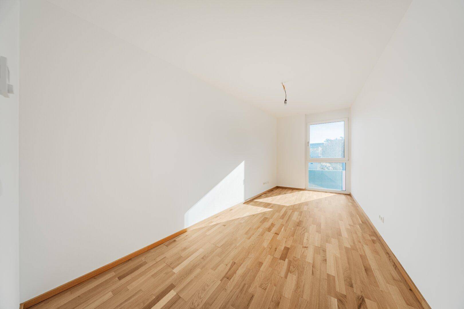 Heller, leerer Raum mit Holzboden und Fenster mit Zugang zu einem kleinen Balkon.