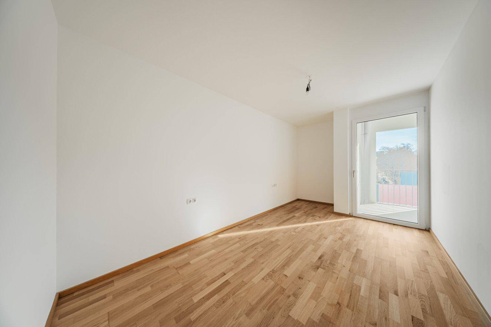 Heller, leerer Raum mit Holzboden und Fenster mit Zugang zu einem kleinen Balkon.