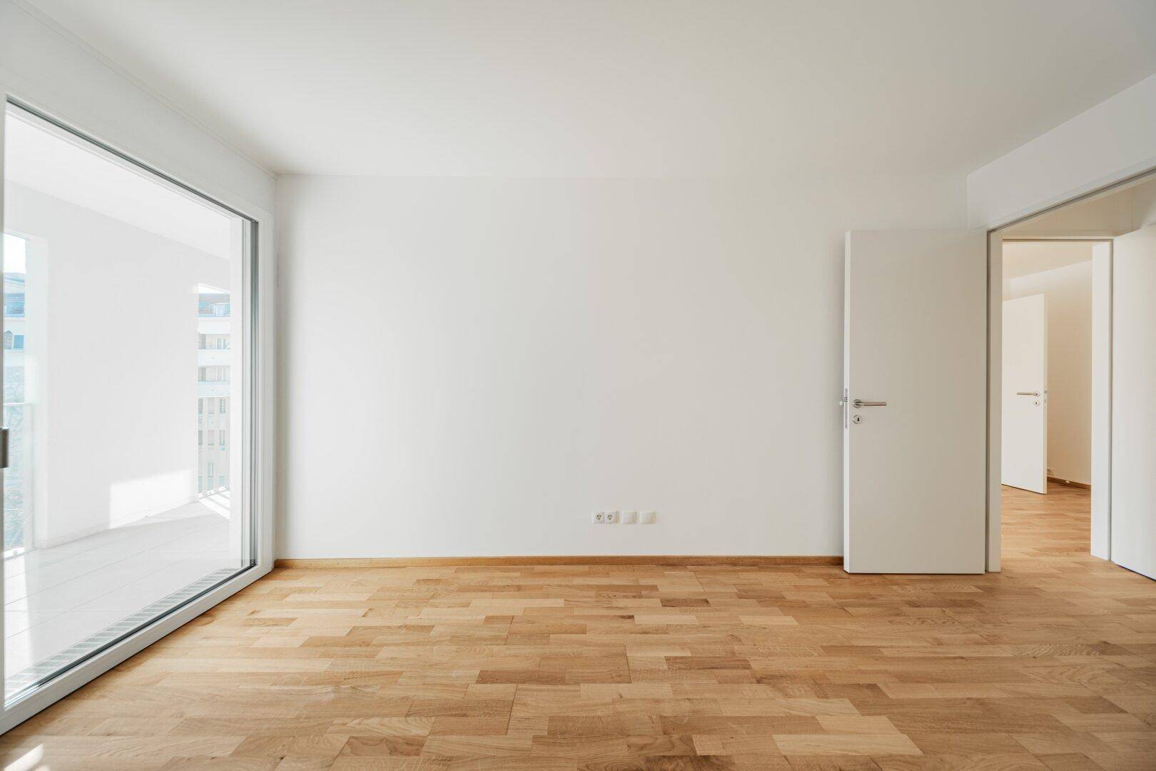 Geräumiges, leeres Zimmer mit Holzboden und Zugang zu einem Balkon.