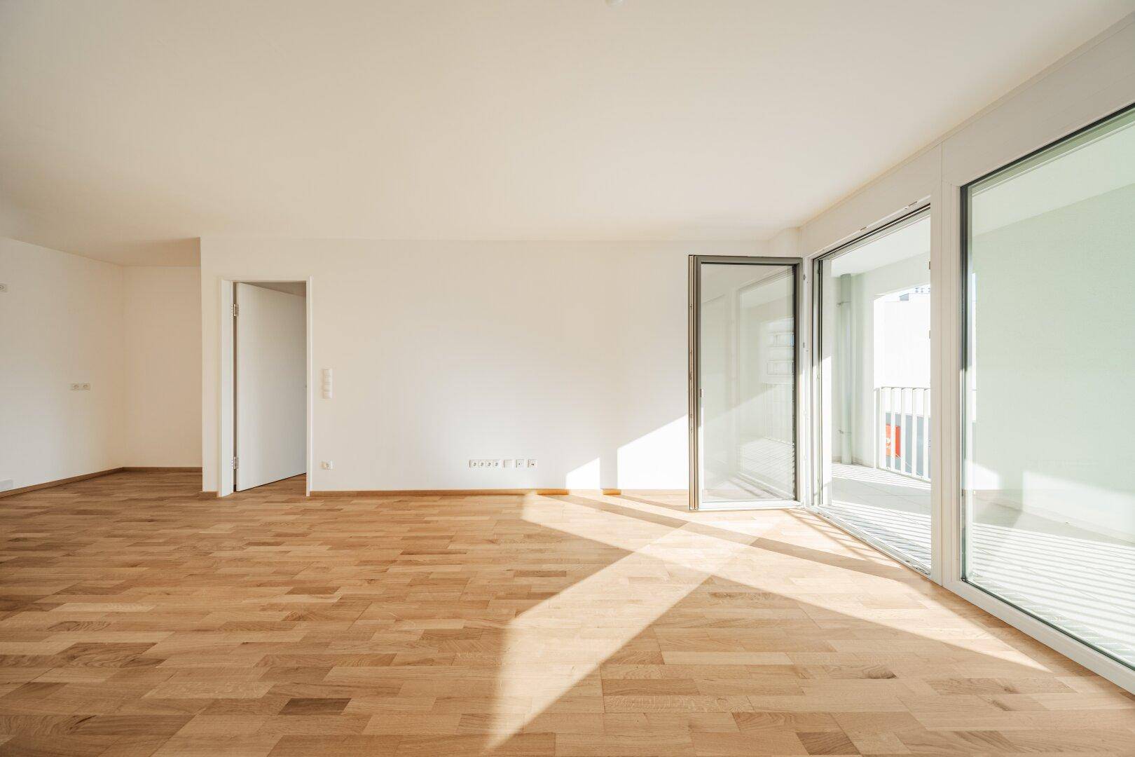 Leeres Wohnzimmer mit Holzboden, großen Fenstern und viel Tageslicht.