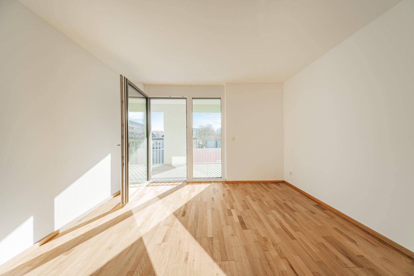 Heller, leerer Raum mit Holzboden und großem Fenster mit Balkonzugang.