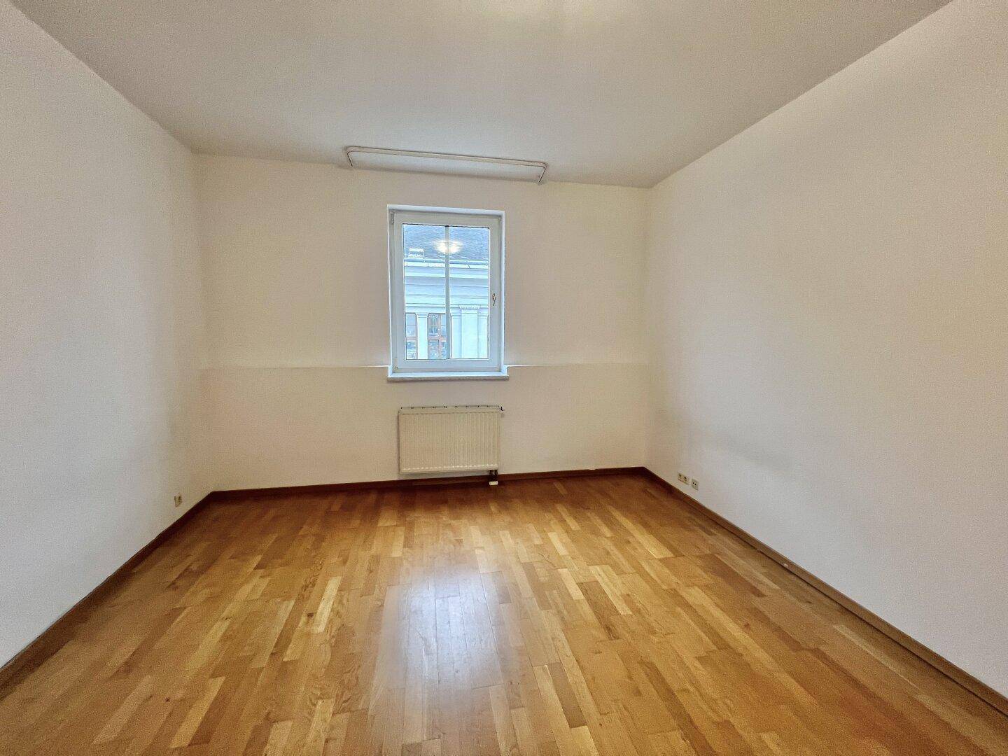 Leeres Zimmer mit hellem Holzfußboden und einem Fenster, ideal als Schlaf- oder Arbeitszimmer.