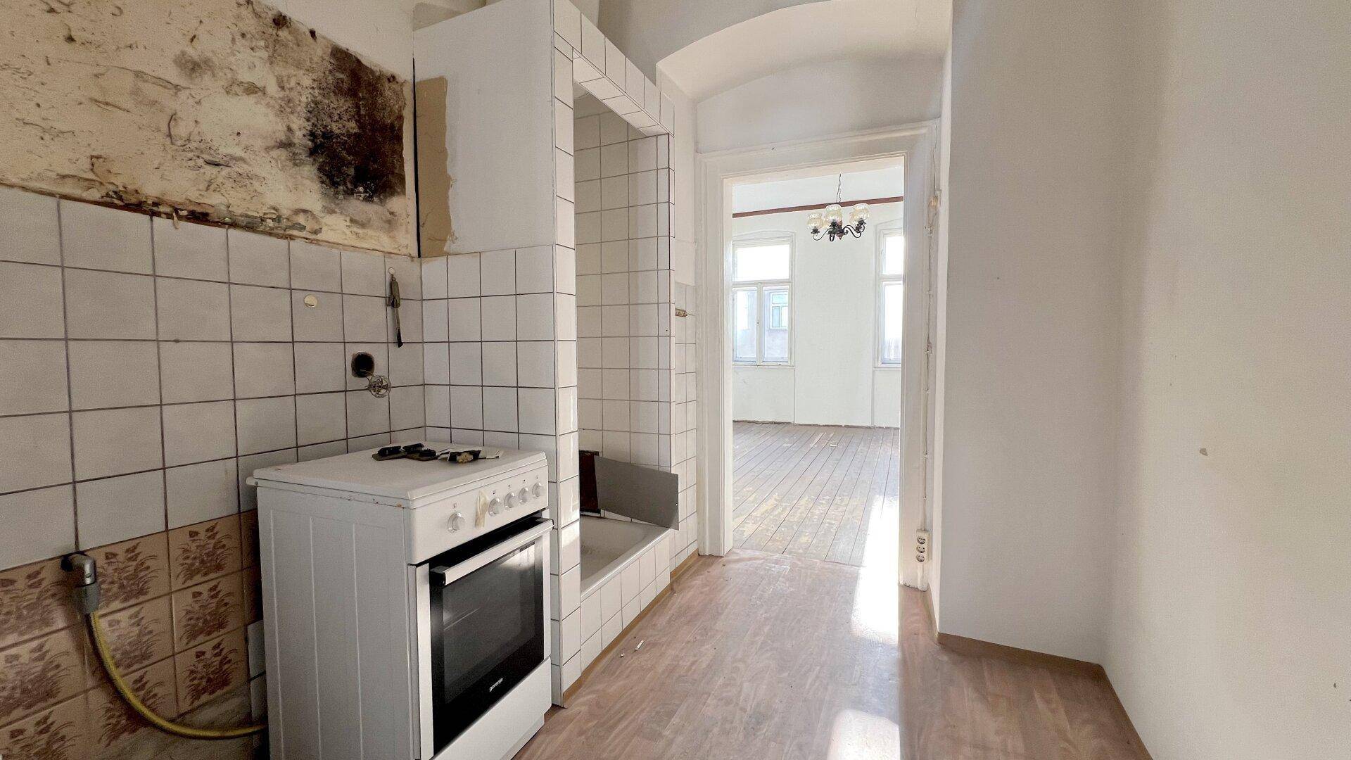 Renovierungsbedürftige Küche mit alten Fliesen, freistehendem Herd und Blick in ein weiteres Zimmer.