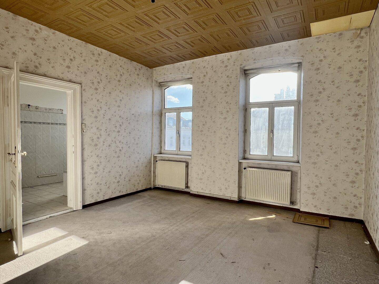 Heller Raum mit zwei Fenstern, gemusterter Tapete und Deckenverkleidung, ideal für eine Renovierung.