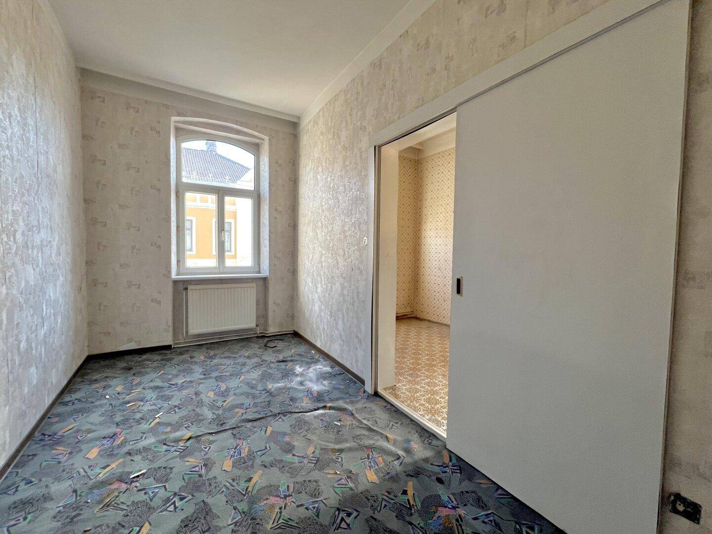 Kleiner Raum mit Schiebetür, gemusterter Tapete und Fenster, ideal als Schlaf- oder Arbeitszimmer.