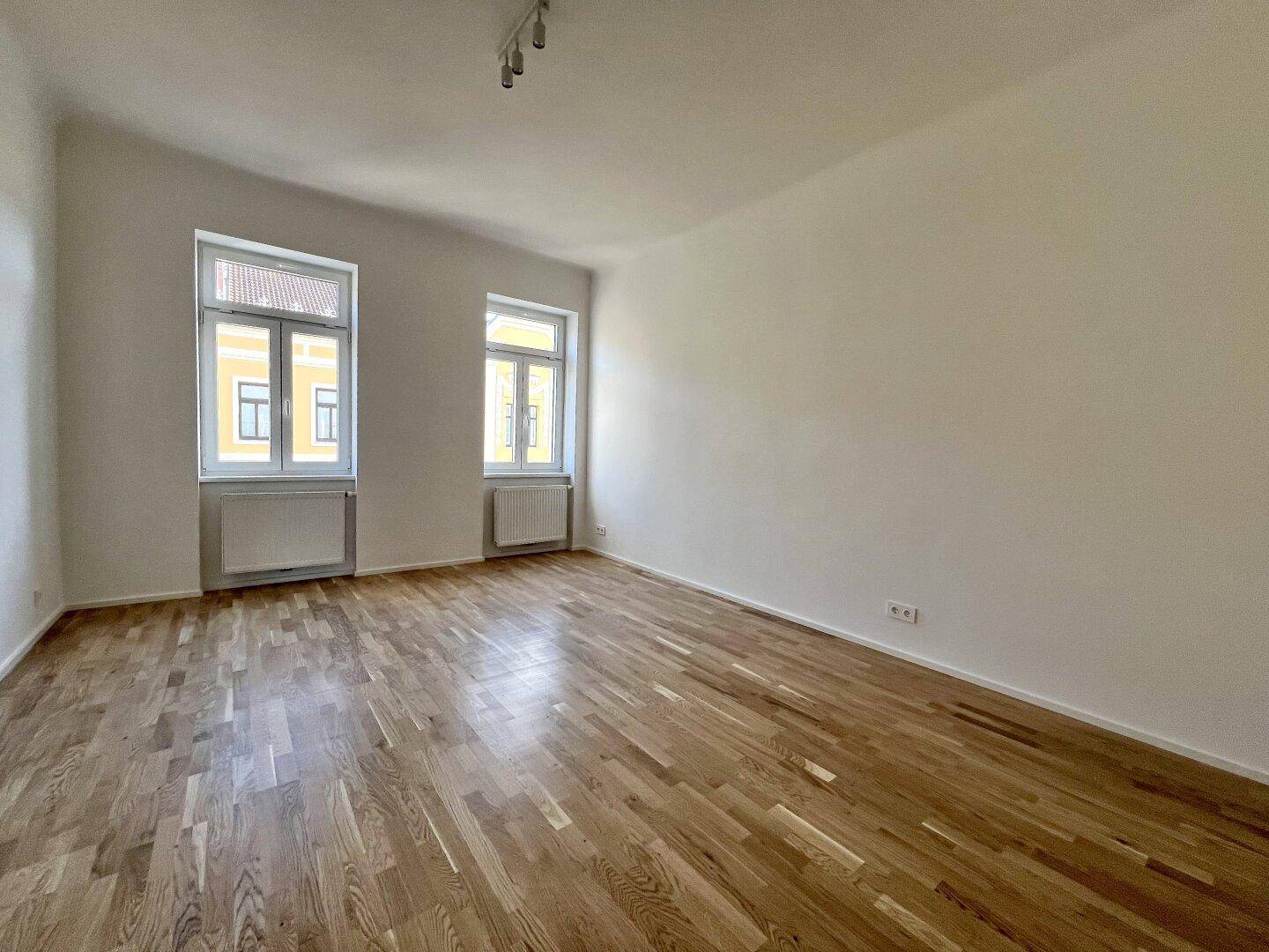 Lichtdurchfluteter Raum mit elegantem Parkettboden und hohen Fenstern, die eine angenehme Atmosphäre schaffen.