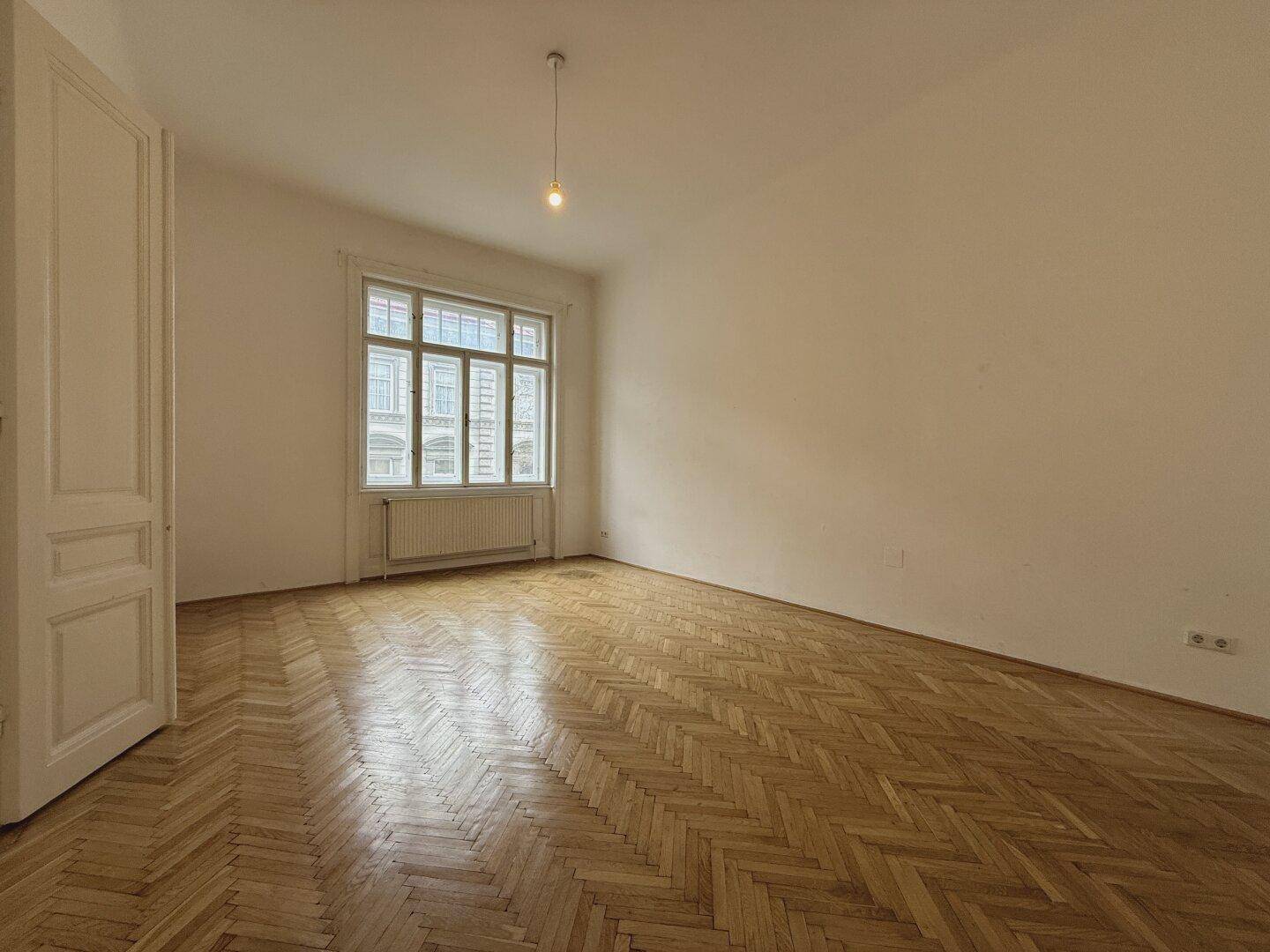 Heller, leerer Raum mit Fischgrätparkett und einem großen Fenster, das viel Tageslicht hereinlässt.