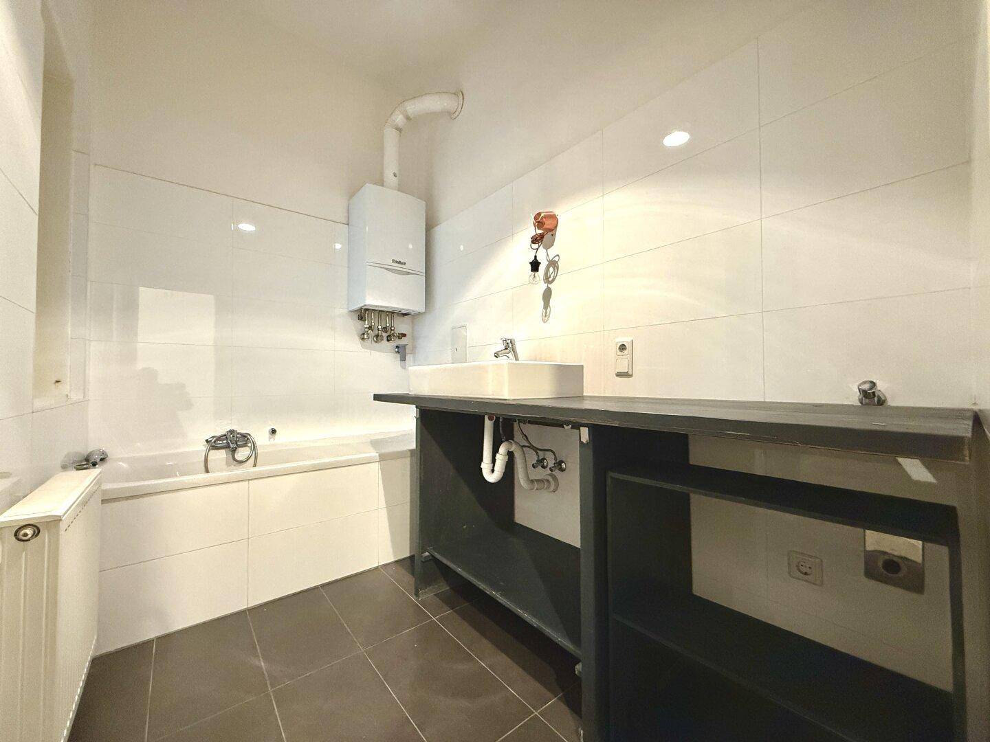 Modern gefliestes Badezimmer mit Badewanne, Waschtisch und einem Warmwasserbereiter an der Wand.