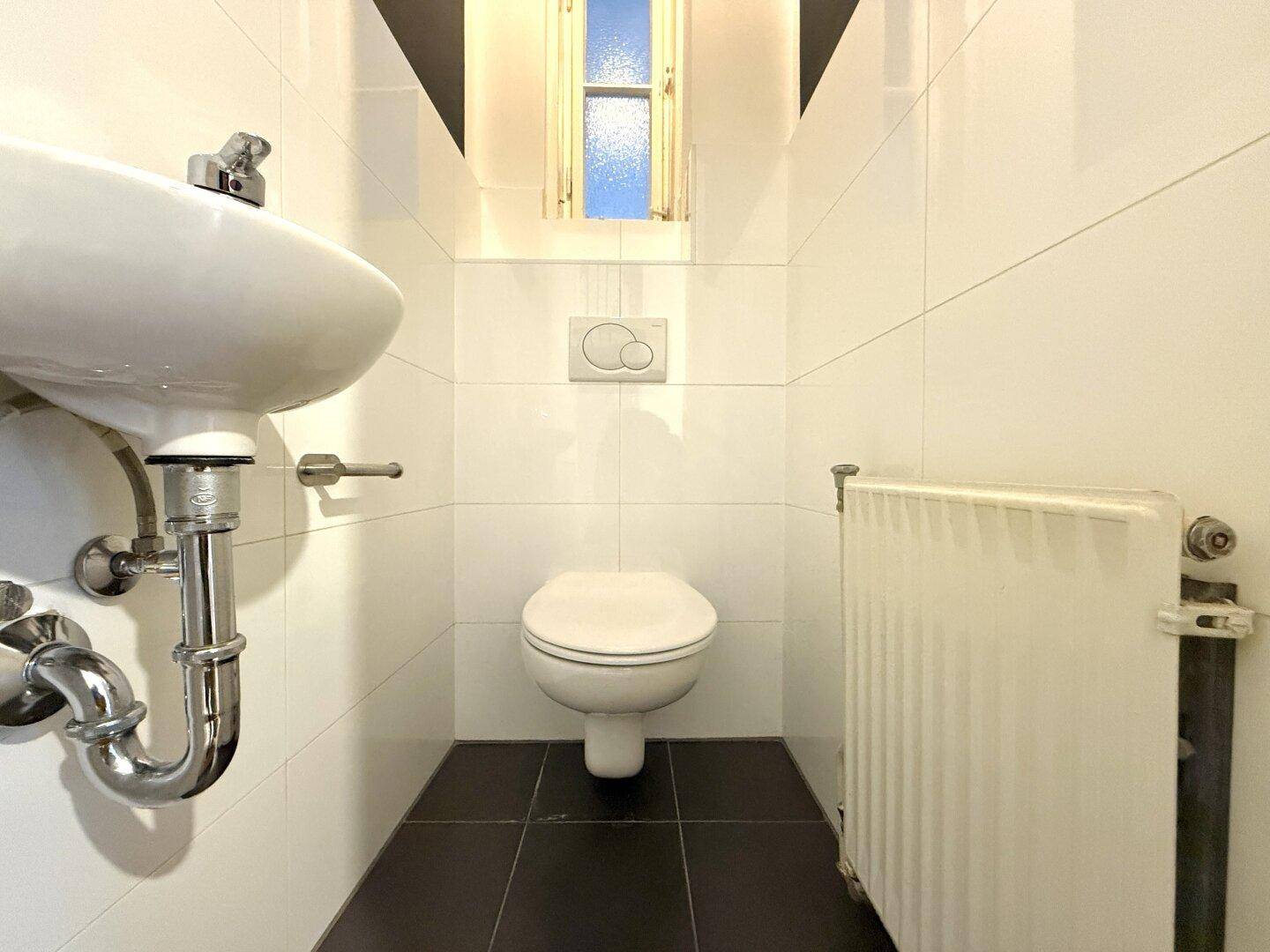 Kleines Gäste-WC mit wandhängender Toilette, Waschbecken, schmalem Fenster und weißen Fliesen.