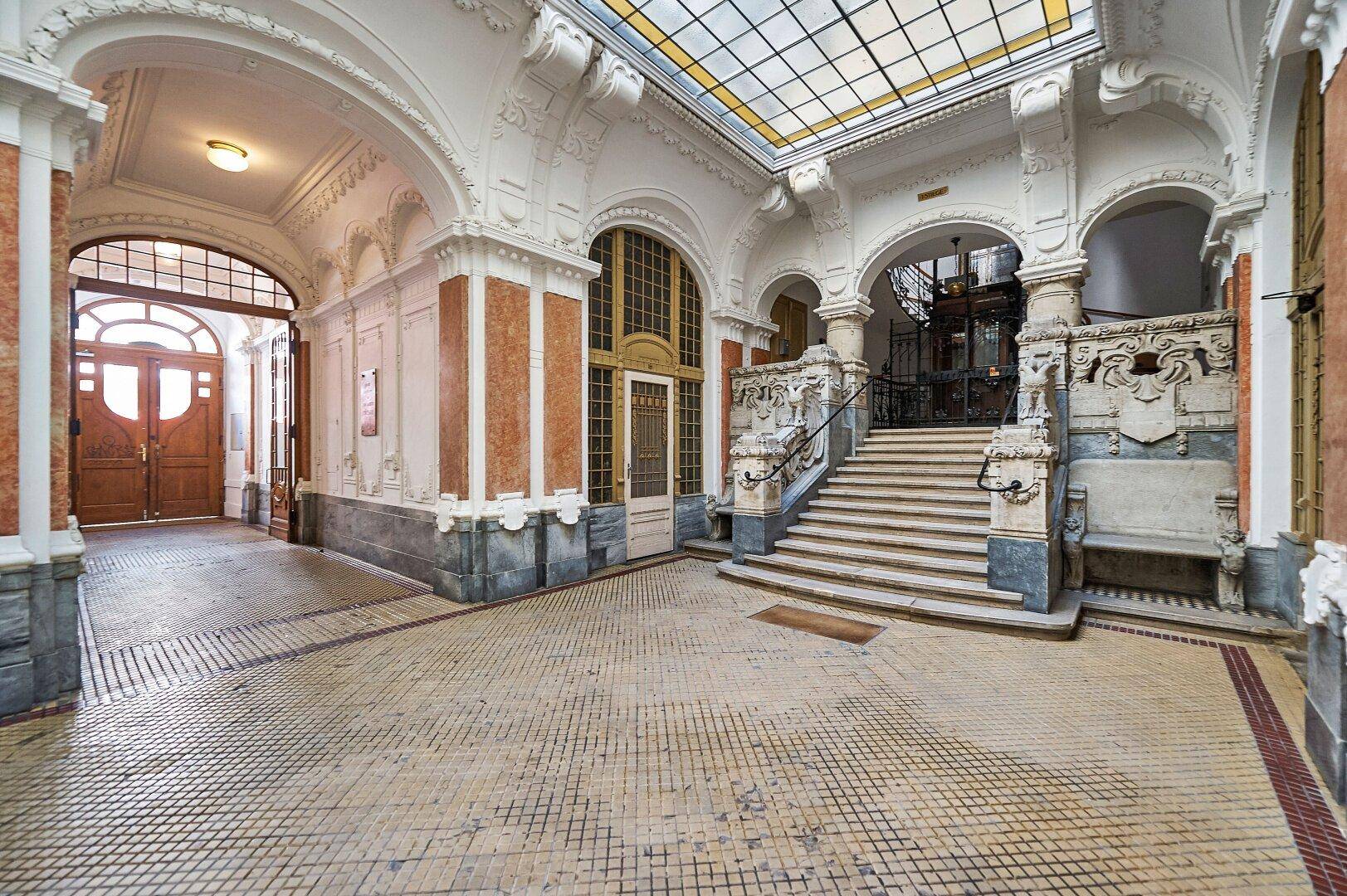 Prunkvolle Eingangshalle eines Altbaus mit beeindruckender Glaskuppel, Marmorverkleidungen und einer breiten Treppe.