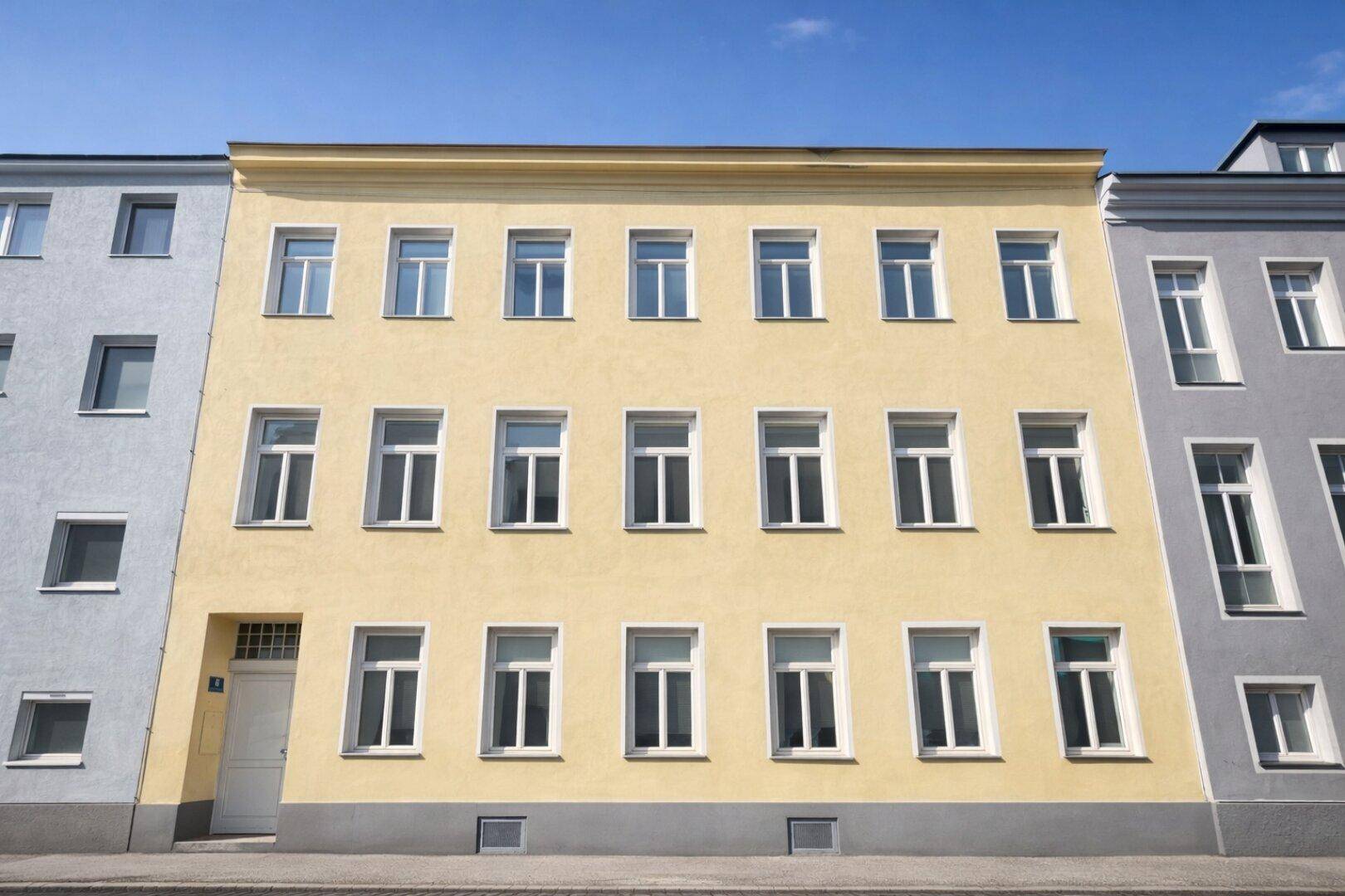 Gelbe Fassade eines Mehrfamilienhauses mit vielen Fenstern unter blauem Himmel.