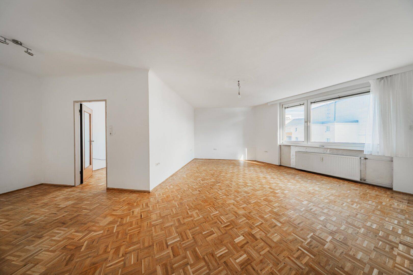Geräumiges, unmöbliertes Wohnzimmer mit Fischgrätparkett und großen Fenstern.
