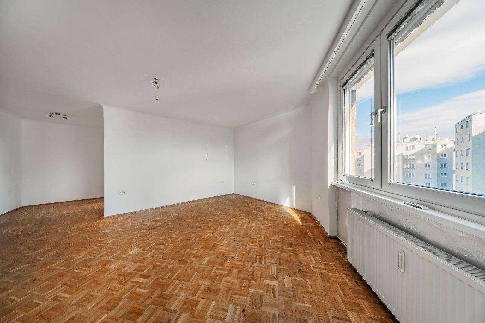 Heller, leerer Raum mit Parkettboden und Blick auf die Stadt durch große Fenster.