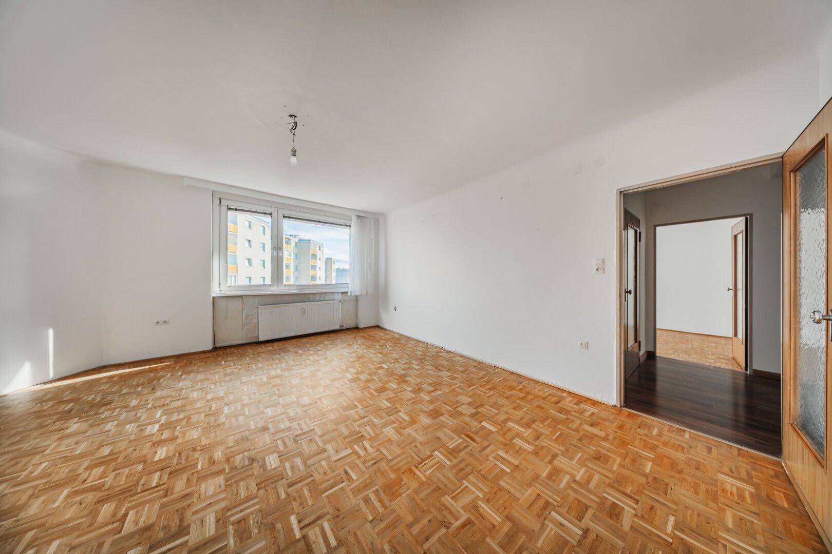 Unmöbliertes Wohnzimmer mit Parkettboden und Zugang zu einem weiteren Raum.