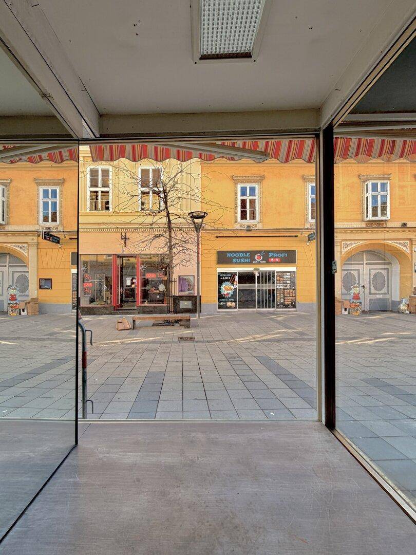 Blick aus einem Ladenlokal auf einen belebten Stadtplatz mit weiteren Geschäften und Gebäuden.
