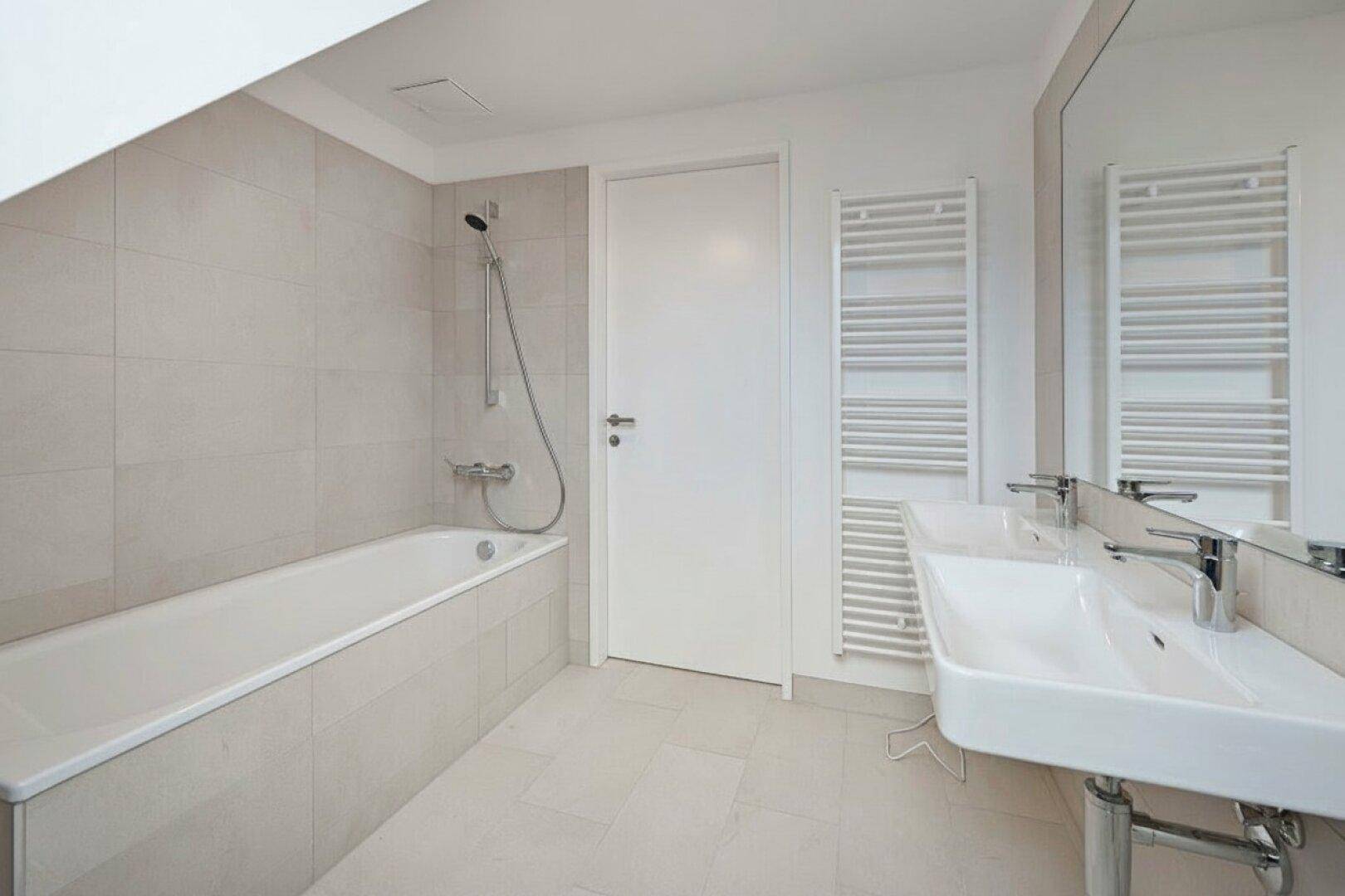 Helles Badezimmer mit Badewanne, Duschfunktion, Einzelwaschbecken und beheiztem Handtuchhalter, modern gefliest.