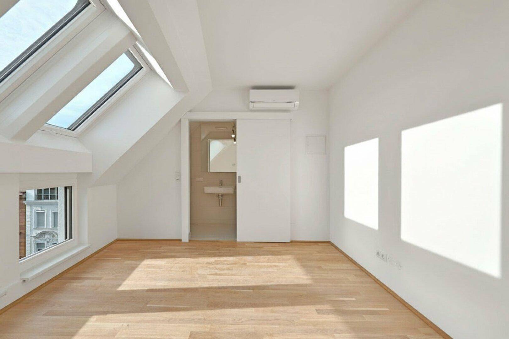Heller Raum mit Holzboden, Dachfenstern und Zugang zu einem Badezimmer durch eine Schiebetür.