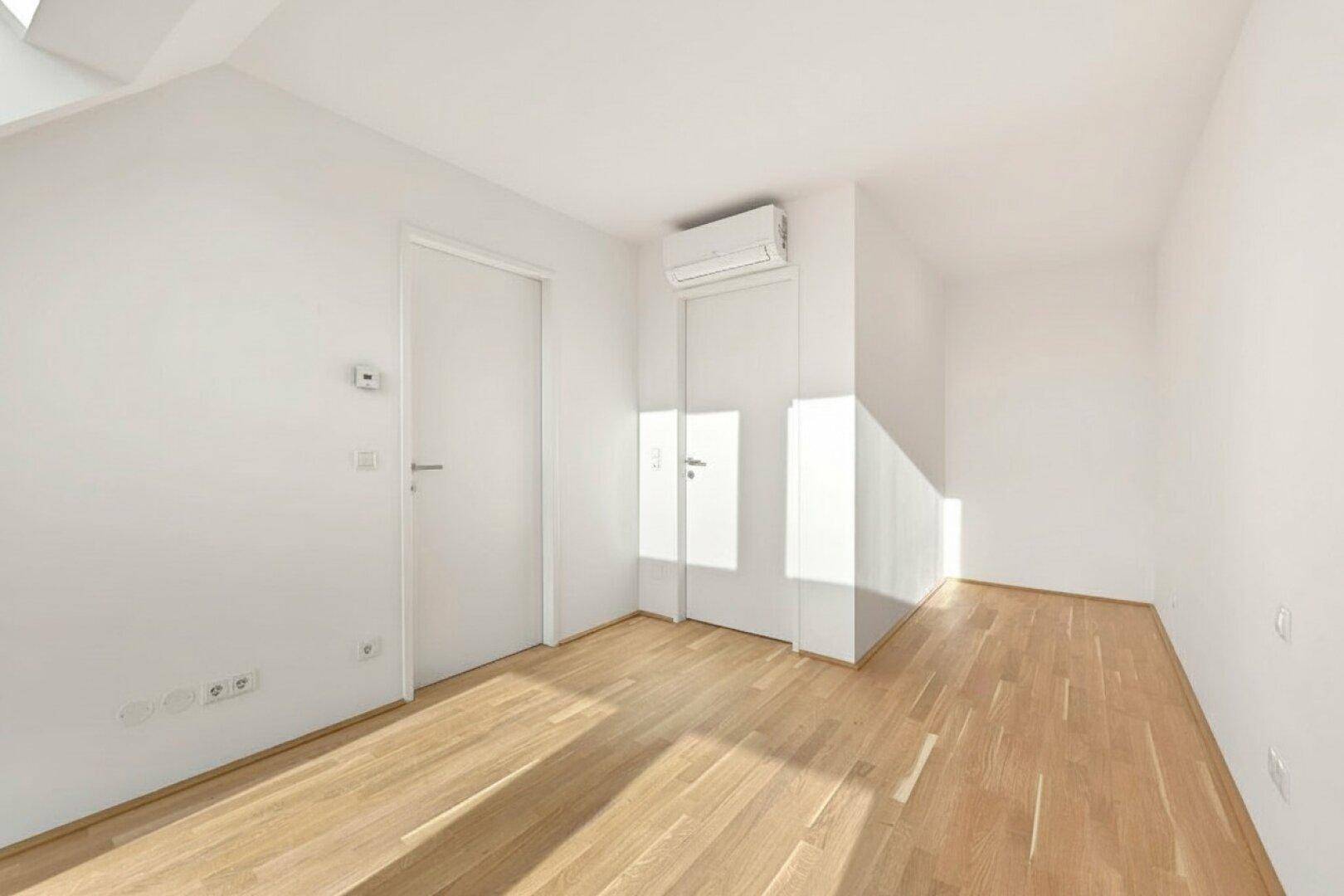 Heller, leerer Raum mit Holzboden, Klimaanlage und weiß gestrichenen Wänden unter Dachschräge.