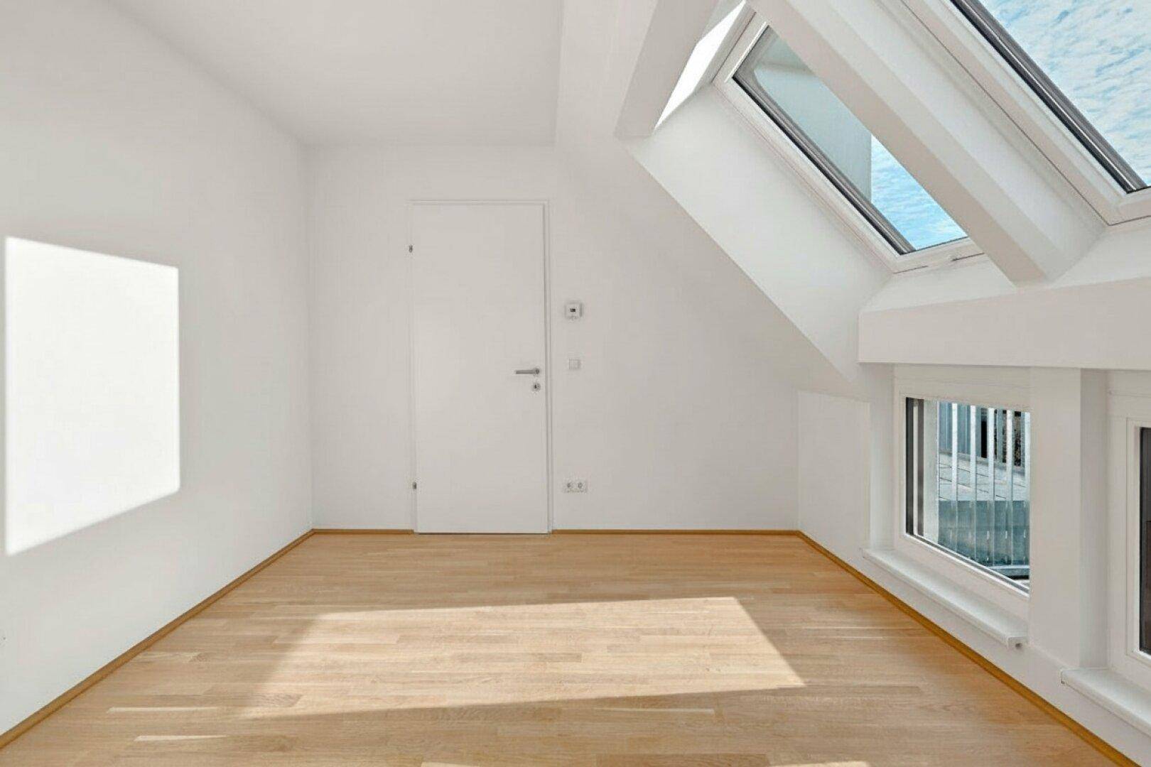 Heller Raum mit Holzboden, Dachfenstern und weiß gestrichenen Wänden unter Dachschräge.