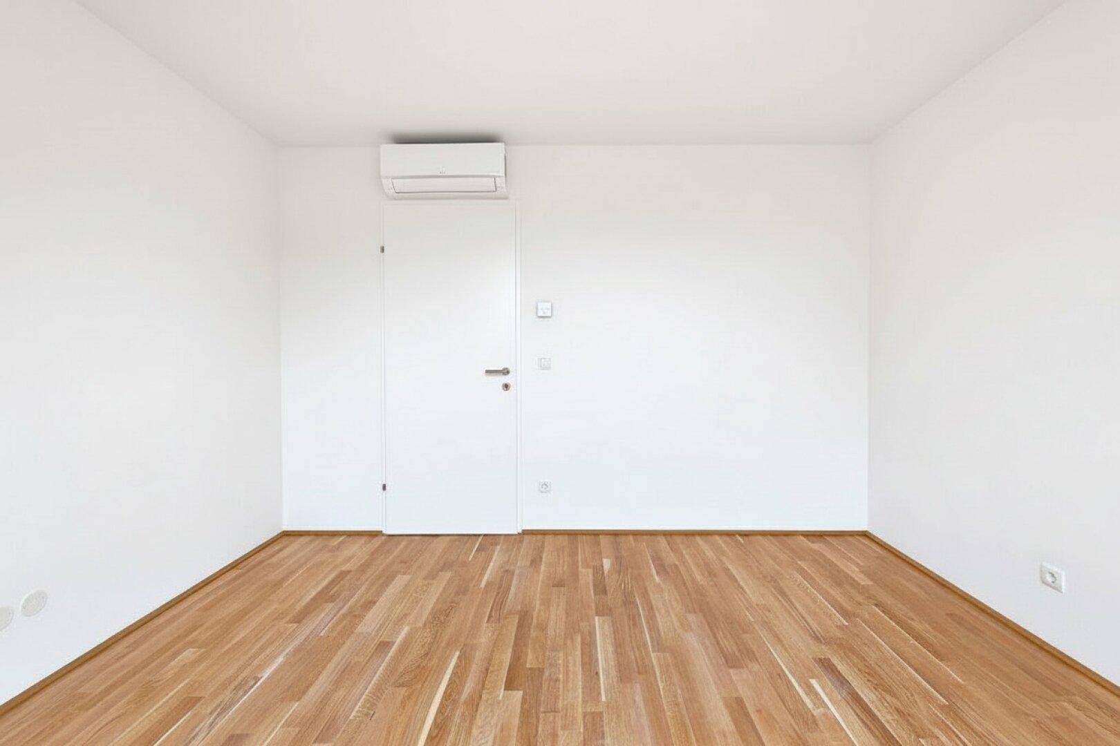 Helles leeres Zimmer mit Holzboden, Klimaanlage und einer weißen Tür.