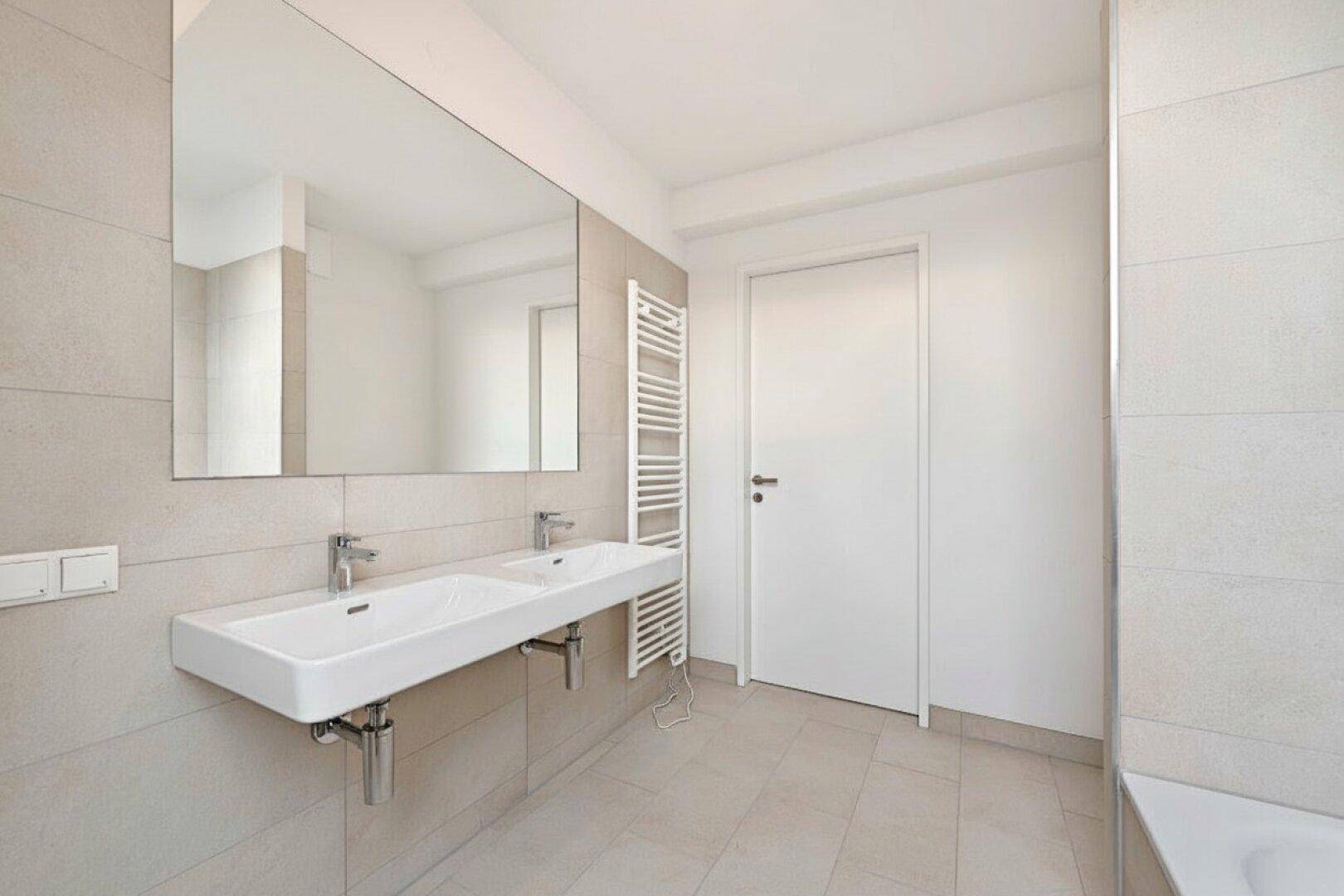 Modernes Badezimmer mit Doppelwaschbecken, großem Spiegel, beheiztem Handtuchhalter und hellen Fliesen.