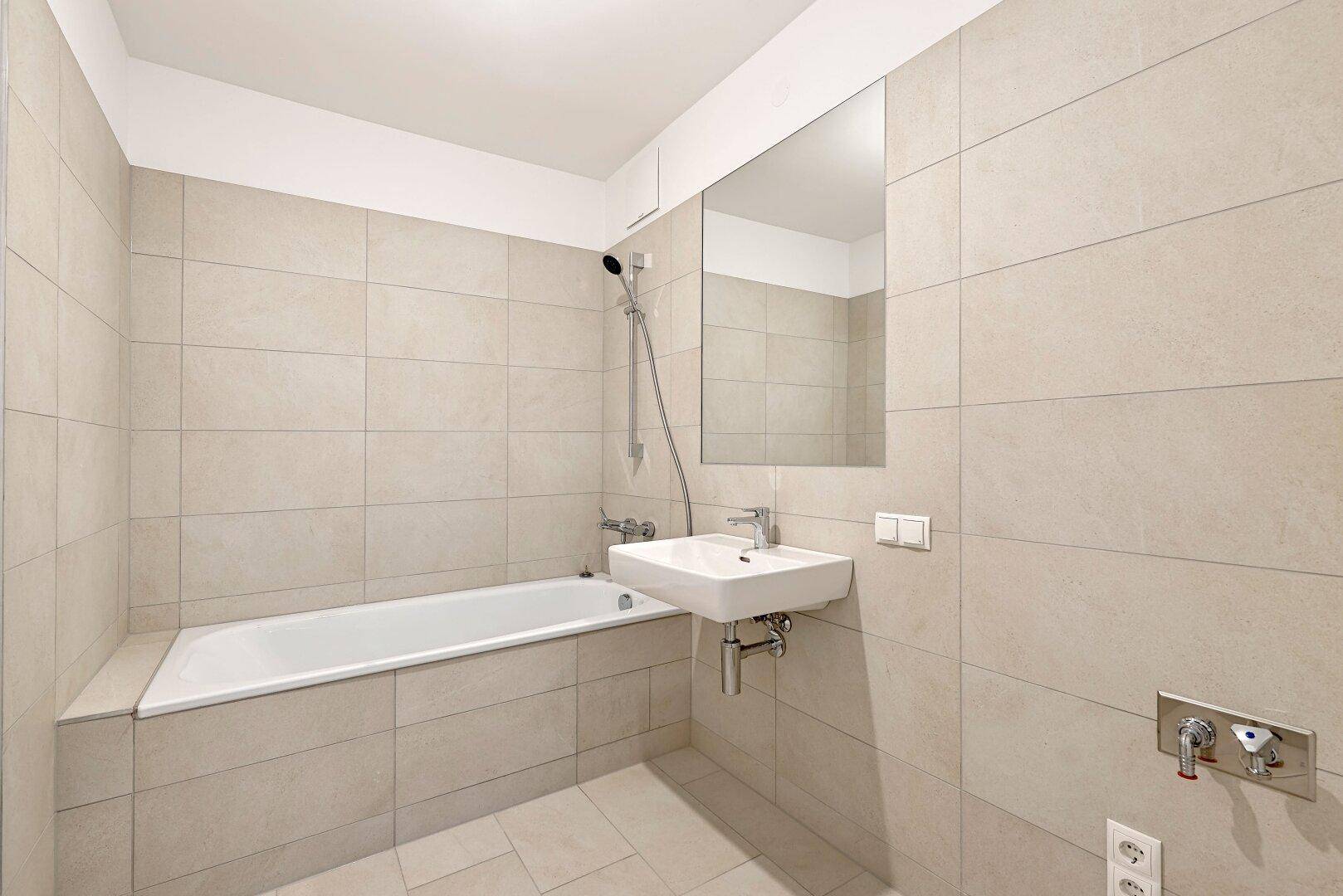 Helles Badezimmer mit Badewanne, Waschbecken und großen, beigefarbenen Wand- und Bodenfliesen.