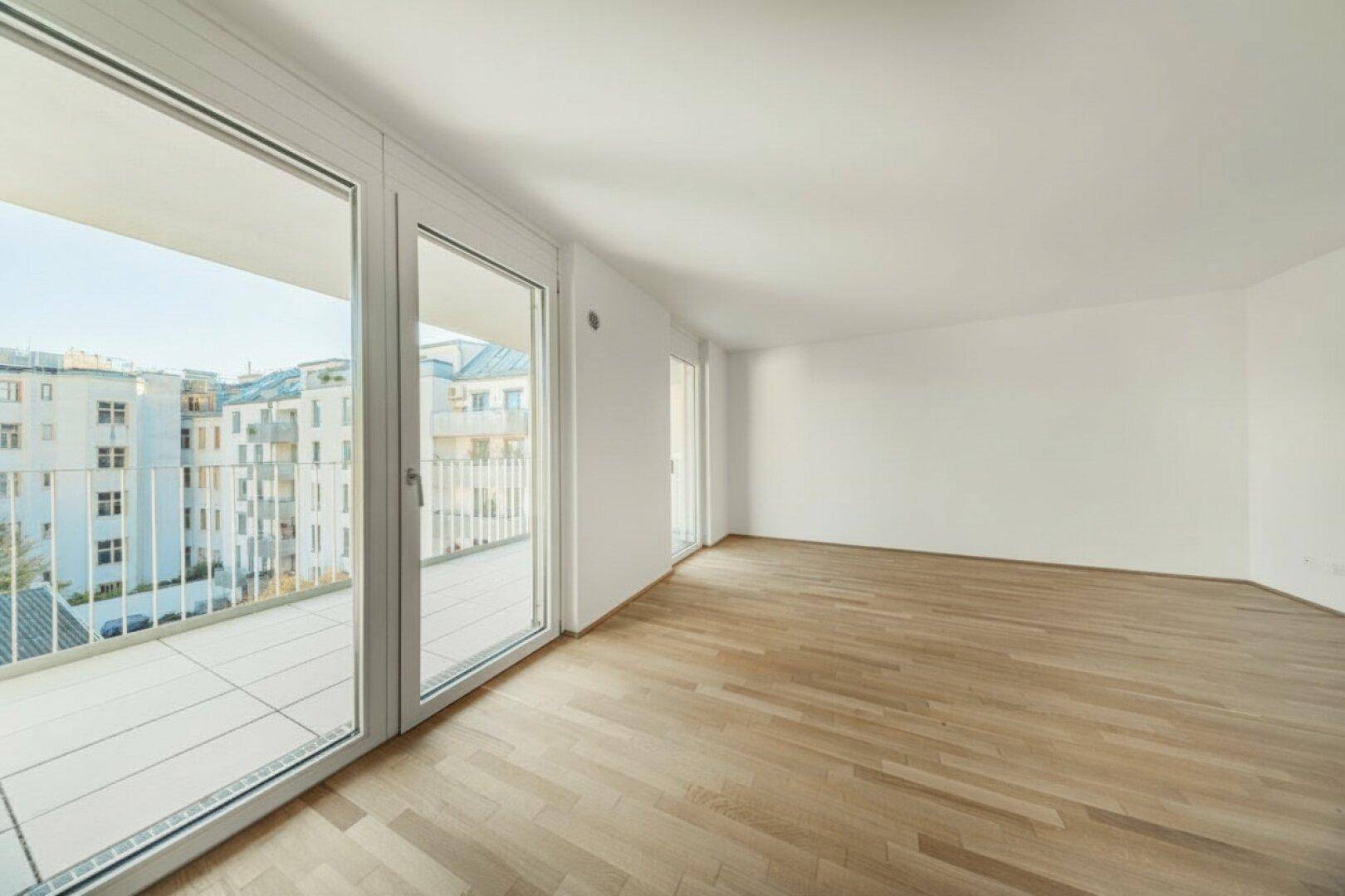 Geräumiges, leeres Wohnzimmer mit hellem Holzboden und Zugang zu einem großen Balkon.