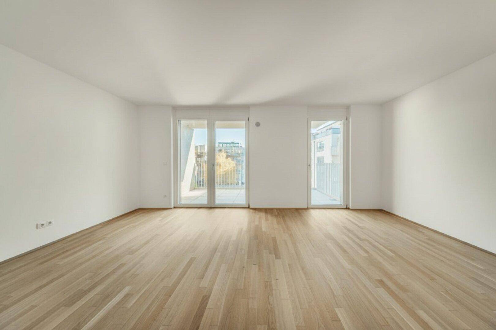 Großes, leeres Wohnzimmer mit Holzboden und zwei Balkonzugängen, sehr hell.