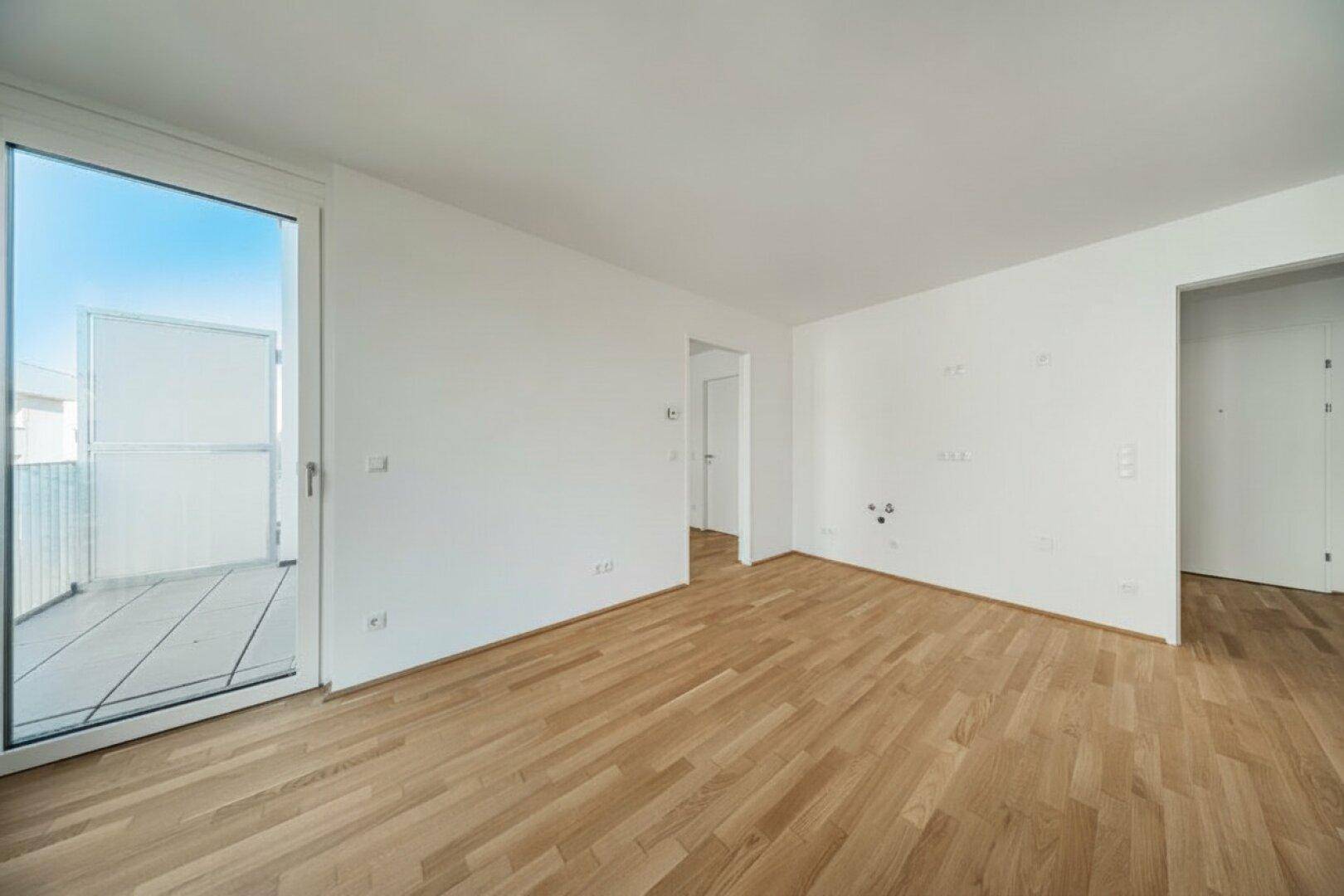 Leerer Raum mit hellem Parkettboden und großem Fenster mit Zugang zu einem Balkon.