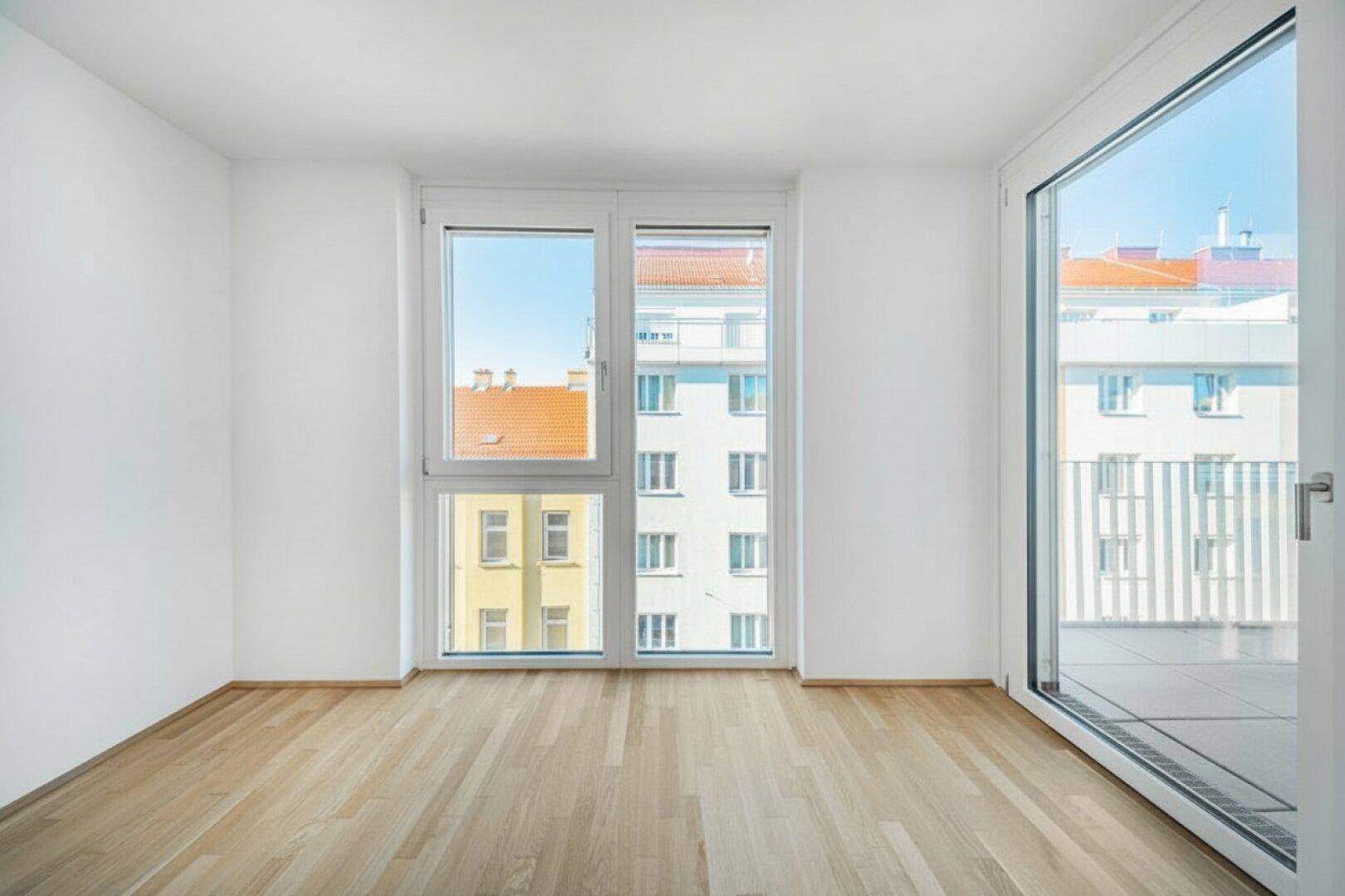 Unmöblierter Raum mit Holzboden, großen Fenstern und Zugang zu einem Außenbereich.