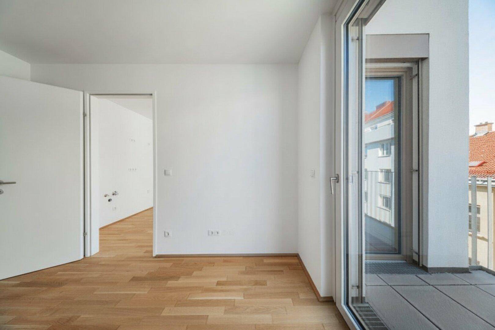 Leerer Raum mit Parkettboden und Fenstertür, die zu einem kleinen Balkon führt.