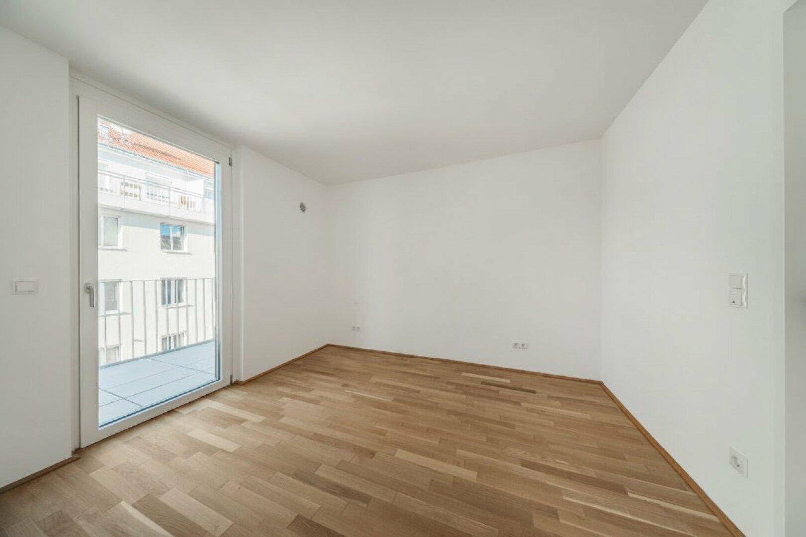 Unmöblierter Raum mit hellem Holzboden und Zugang zu einem Balkon mit Stadtblick.