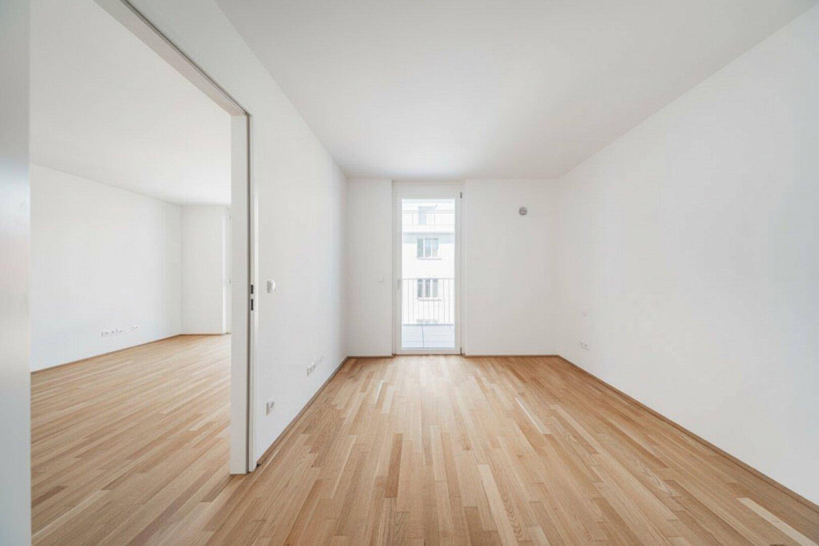 Heller Raum mit Parkettboden und Fenstertür, die zu einem französischen Balkon führt.